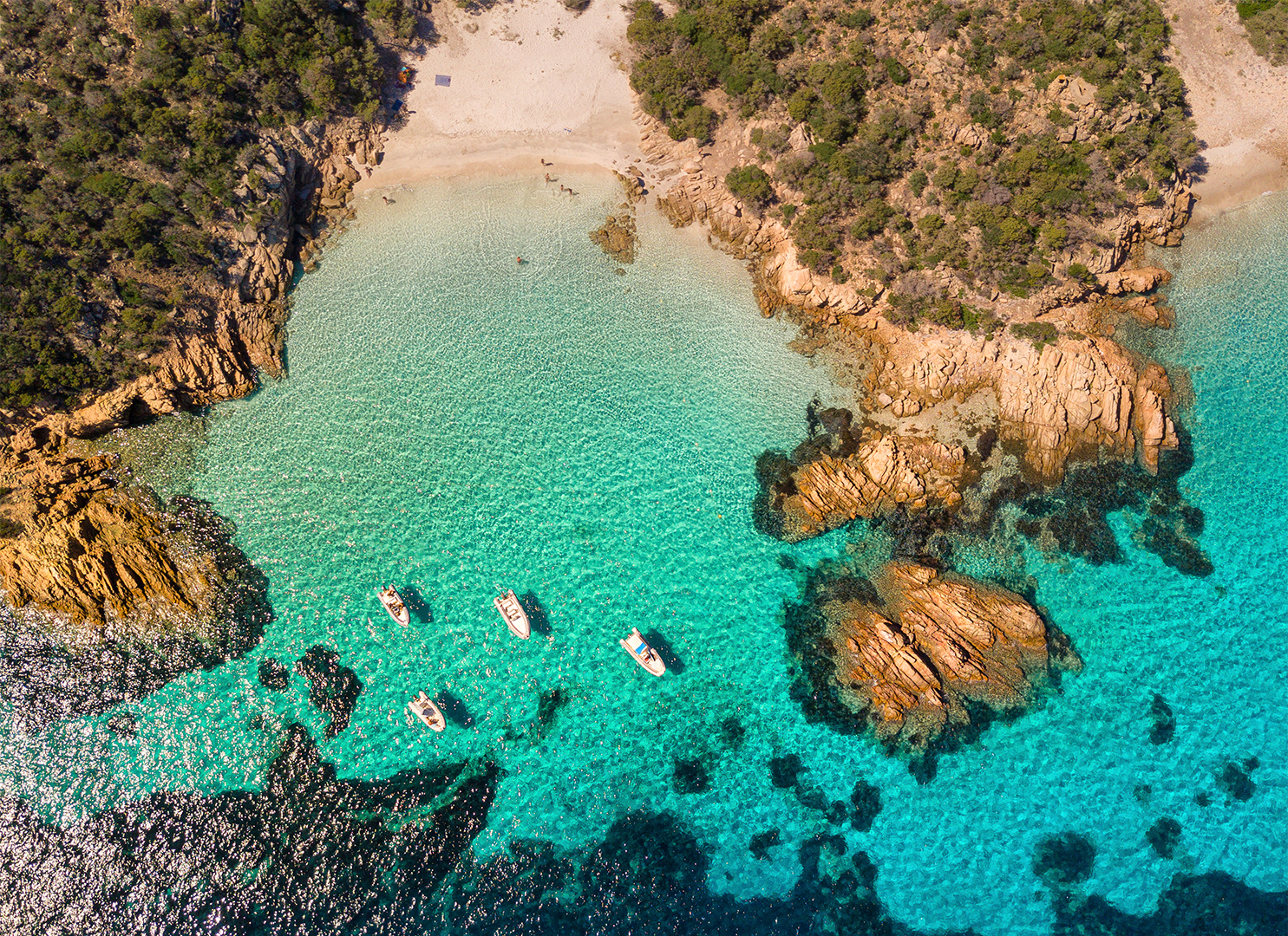 Aerial view of a clear turquoise sea with boats near the shore, surrounded by rocky outcrops and trees.