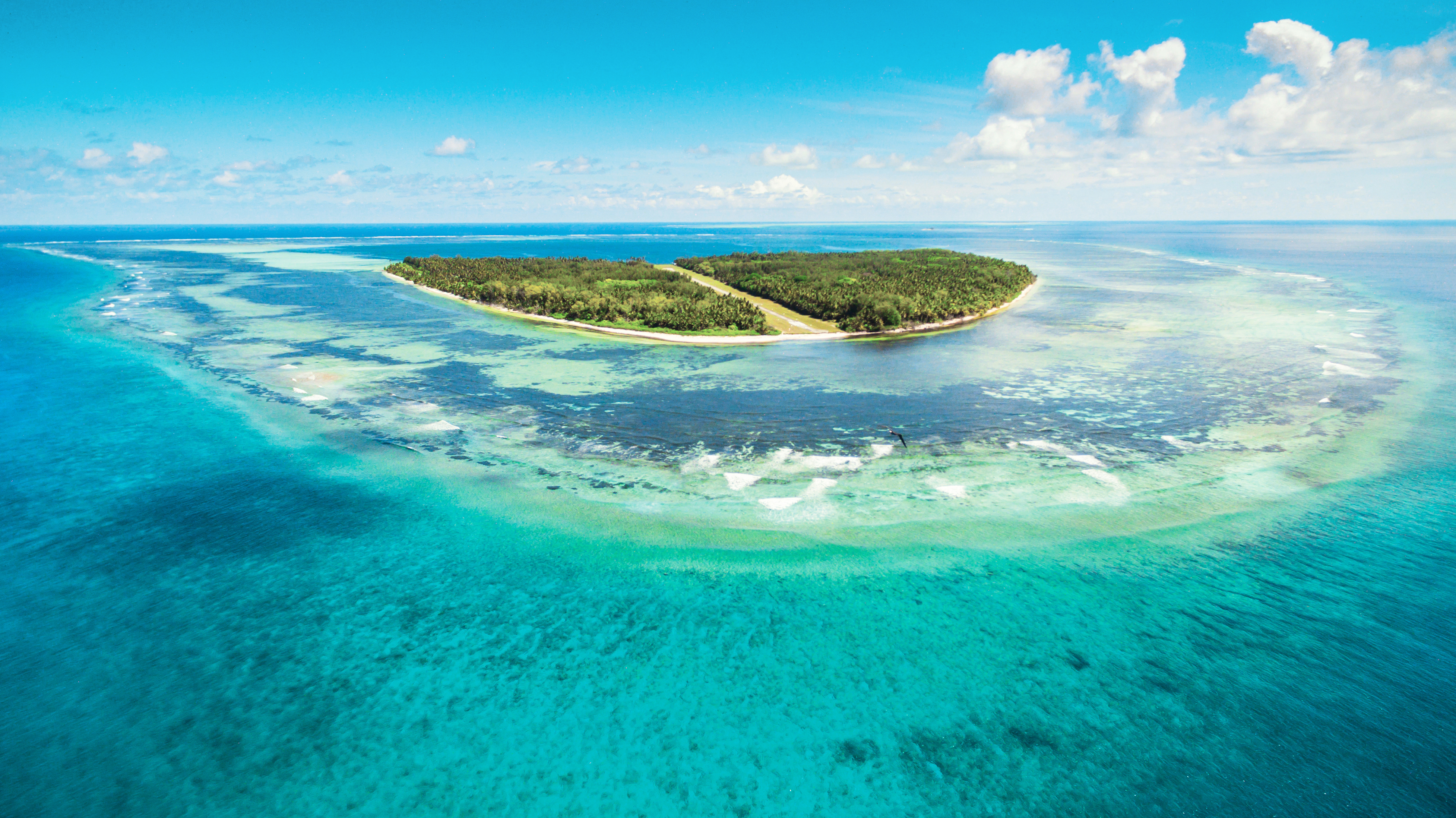 A vast pcean landscape with a lush green island surrounded by reefs in the centre