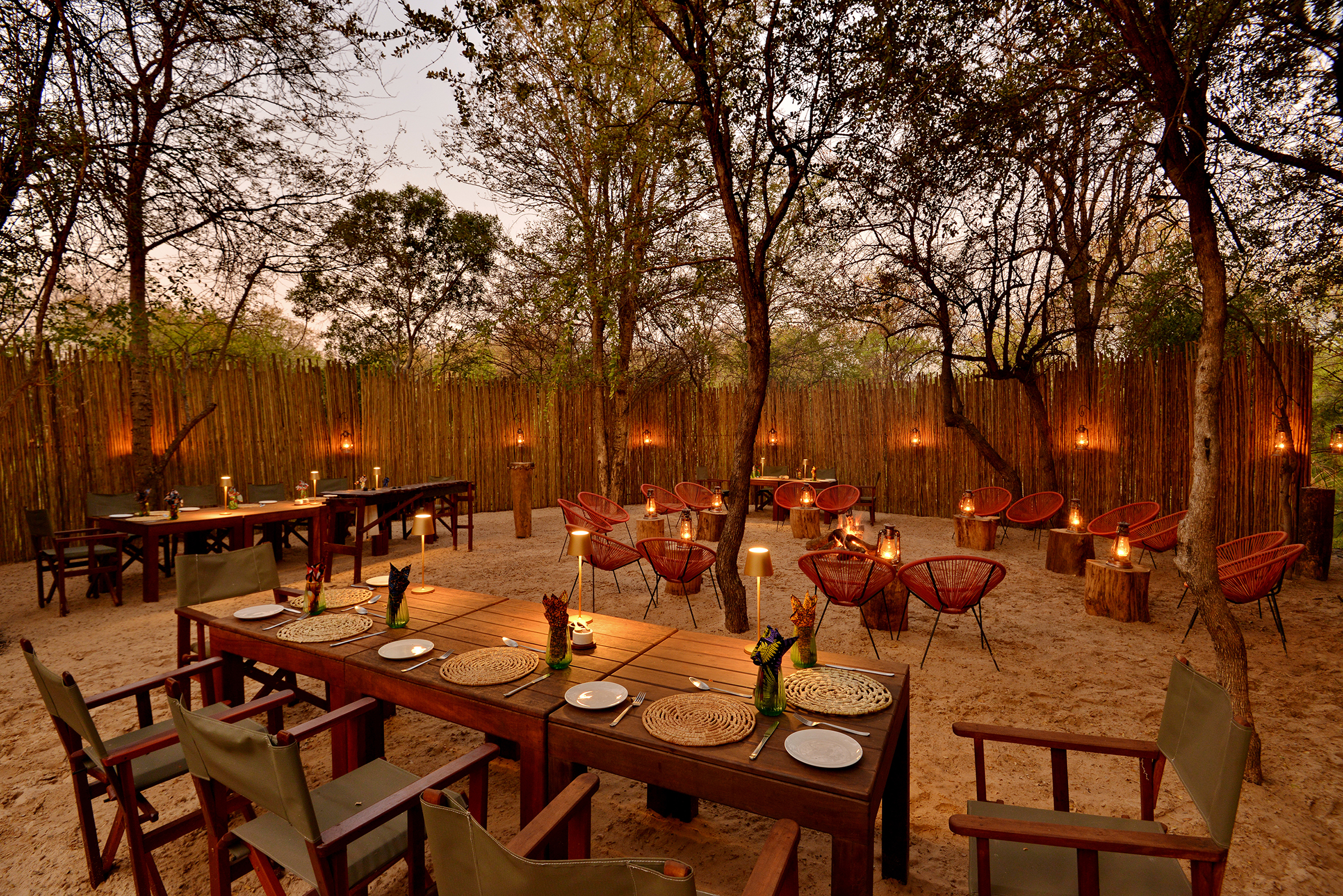 An outdoor Boma dining area at Mukwa River Lodge with wooden tables and chairs, surrounded by trees and lit lanterns, creating a cozy and inviting atmosphere.