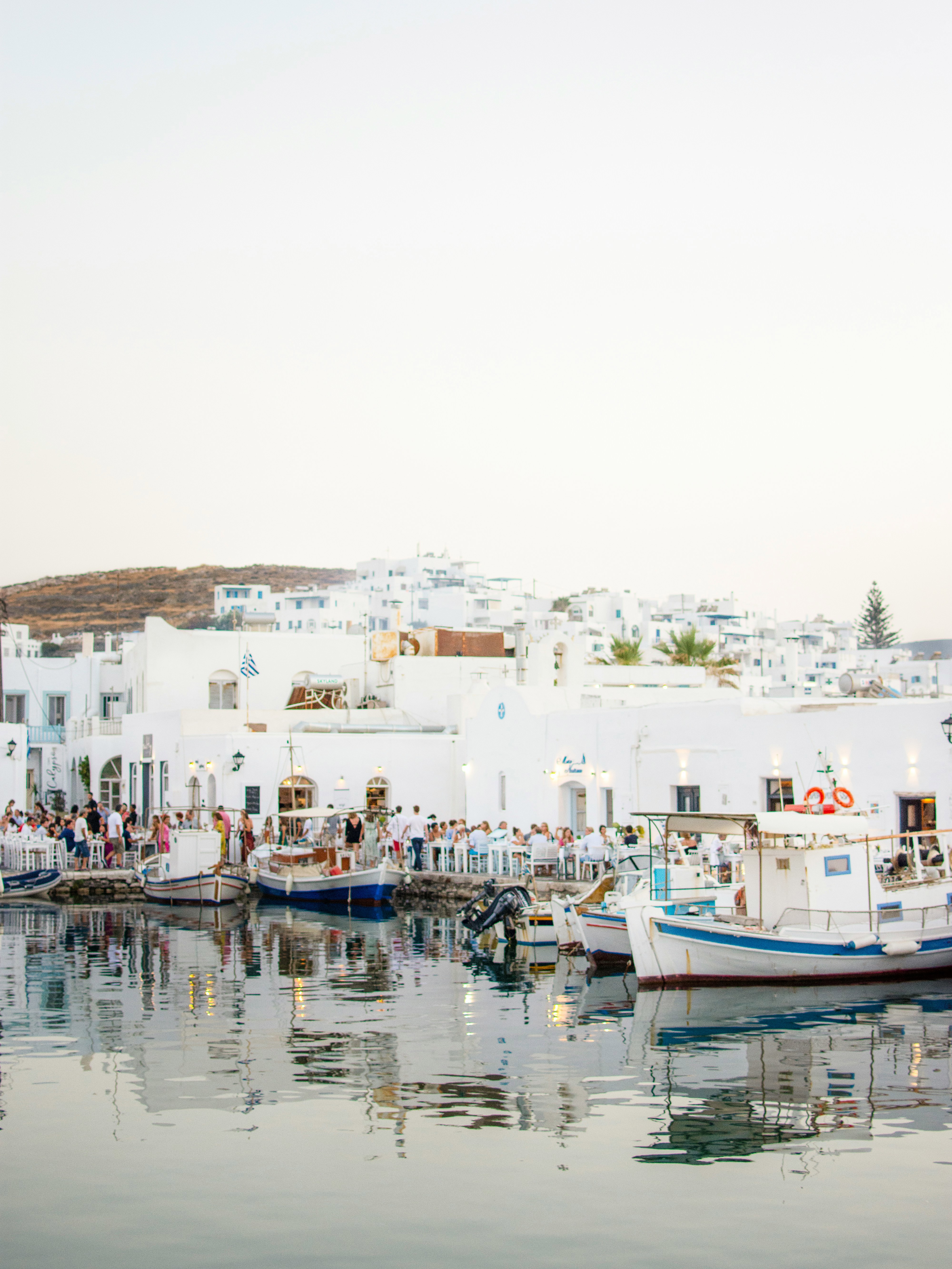 View across the Naoussa marina of people dining by the water with boats moored up at the side