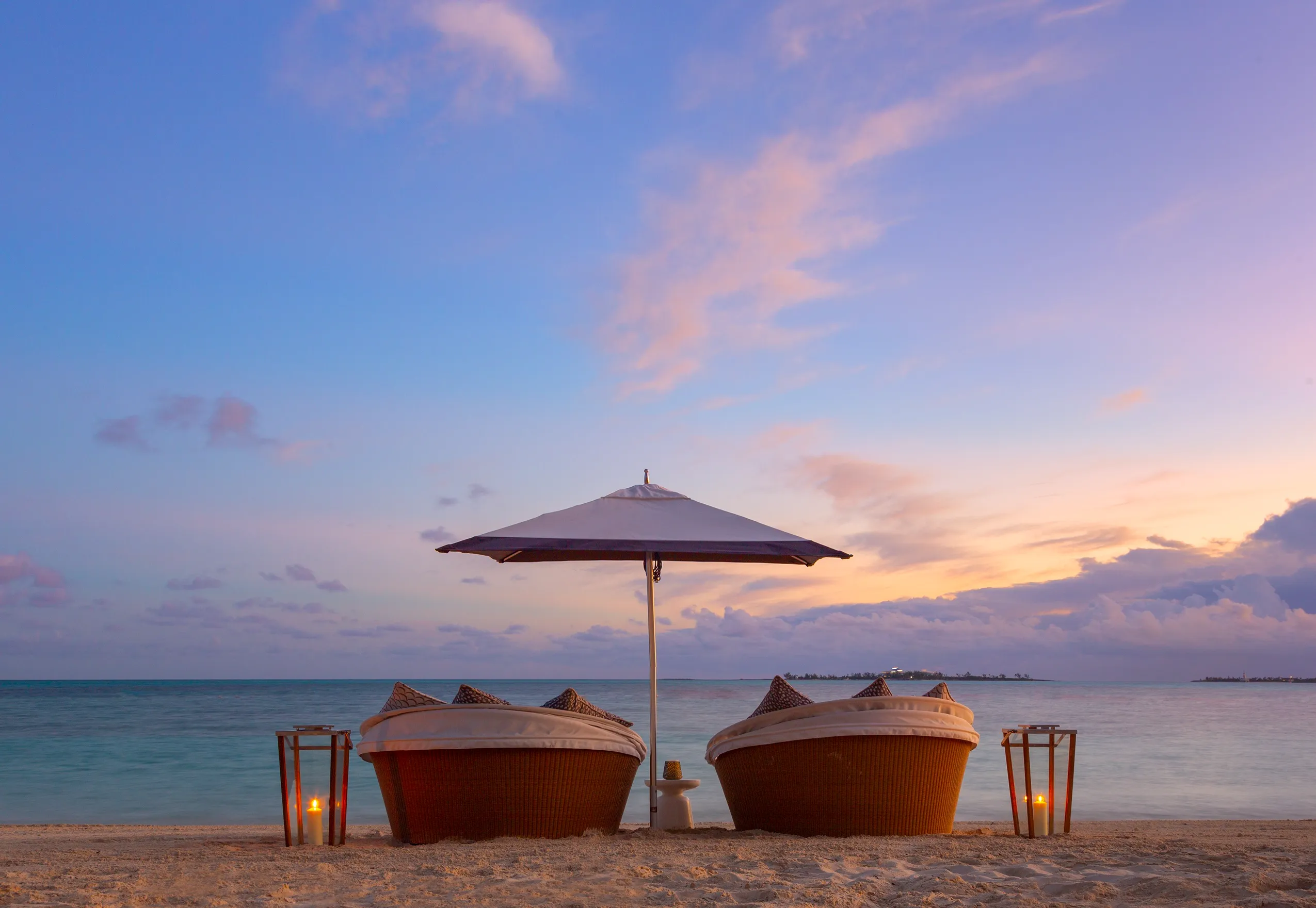 Two wicker lounge chairs under a white umbrella on a sandy beach at sunset