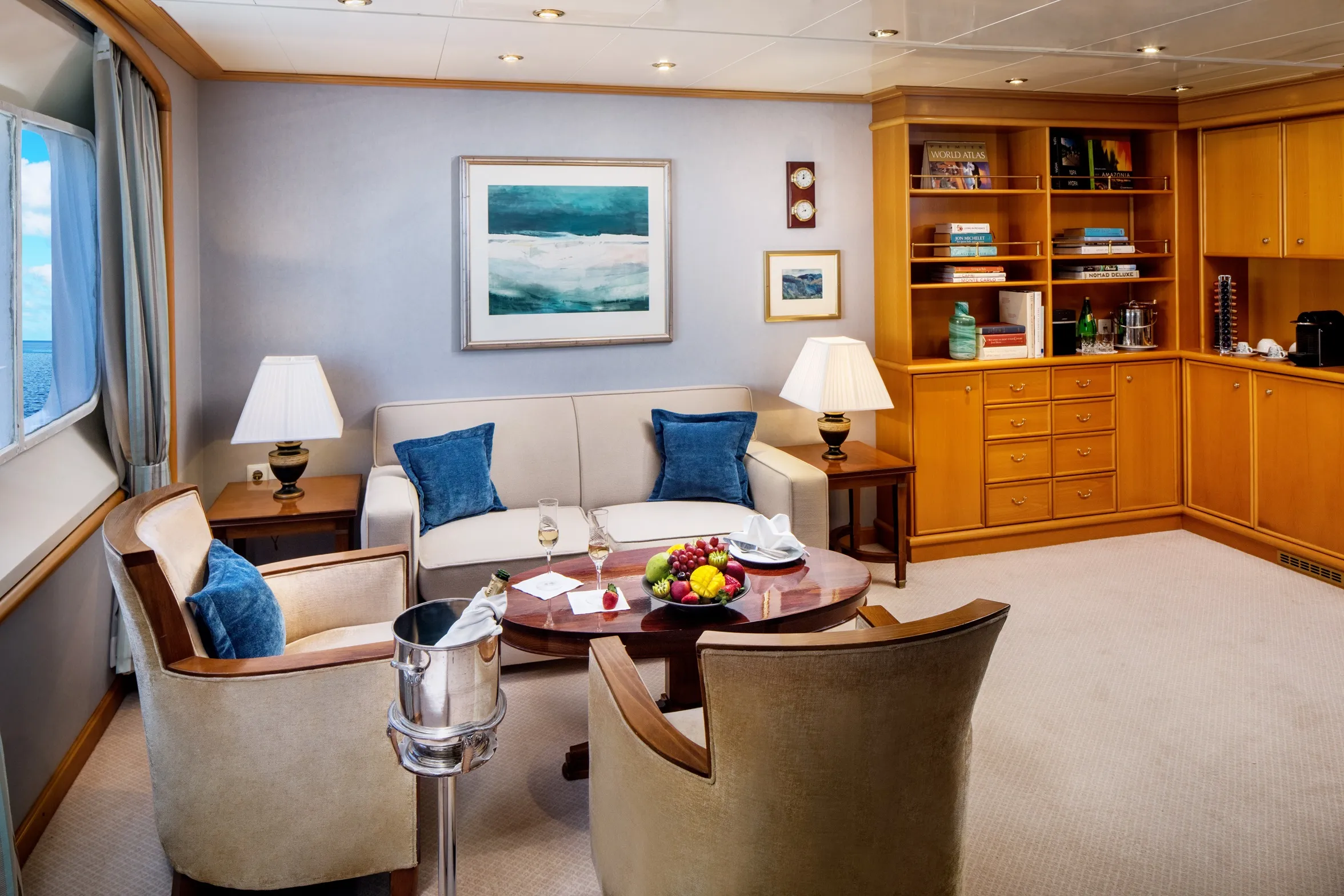 Luxurious living room with beige sofa, blue cushions, armchairs, and a round wooden table with fruit and drinks.