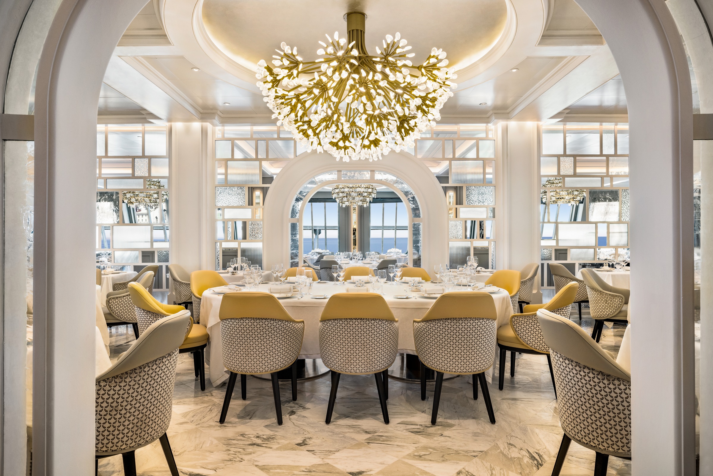 Luxurious Oceania Cruises dining room with a round table, upholstered chairs, ornate chandelier, and arched mirrors.