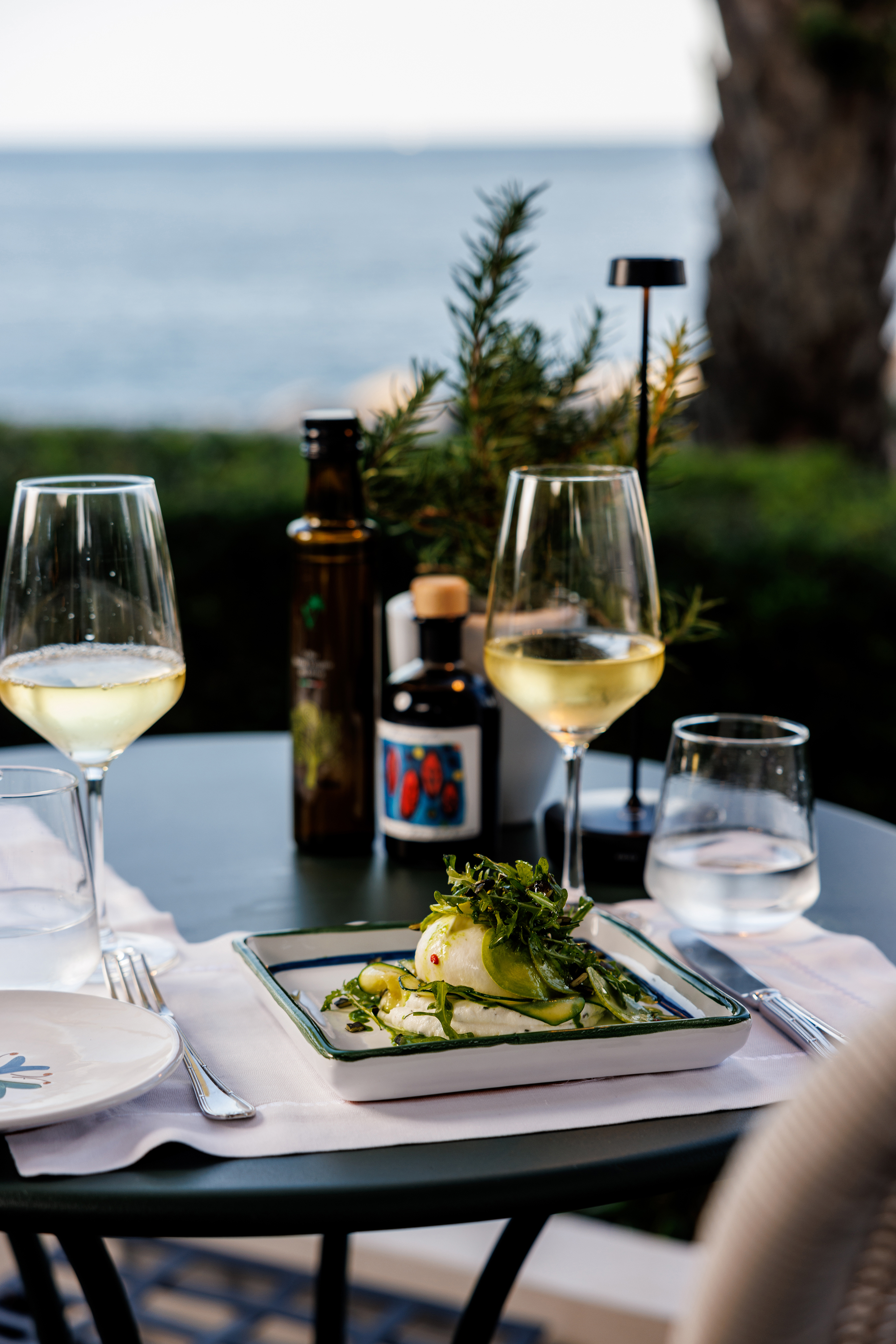 A dining table set with two glasses of white wine, a bottle of olive oil, and a plate of food with greens, overlooking a serene sea view.