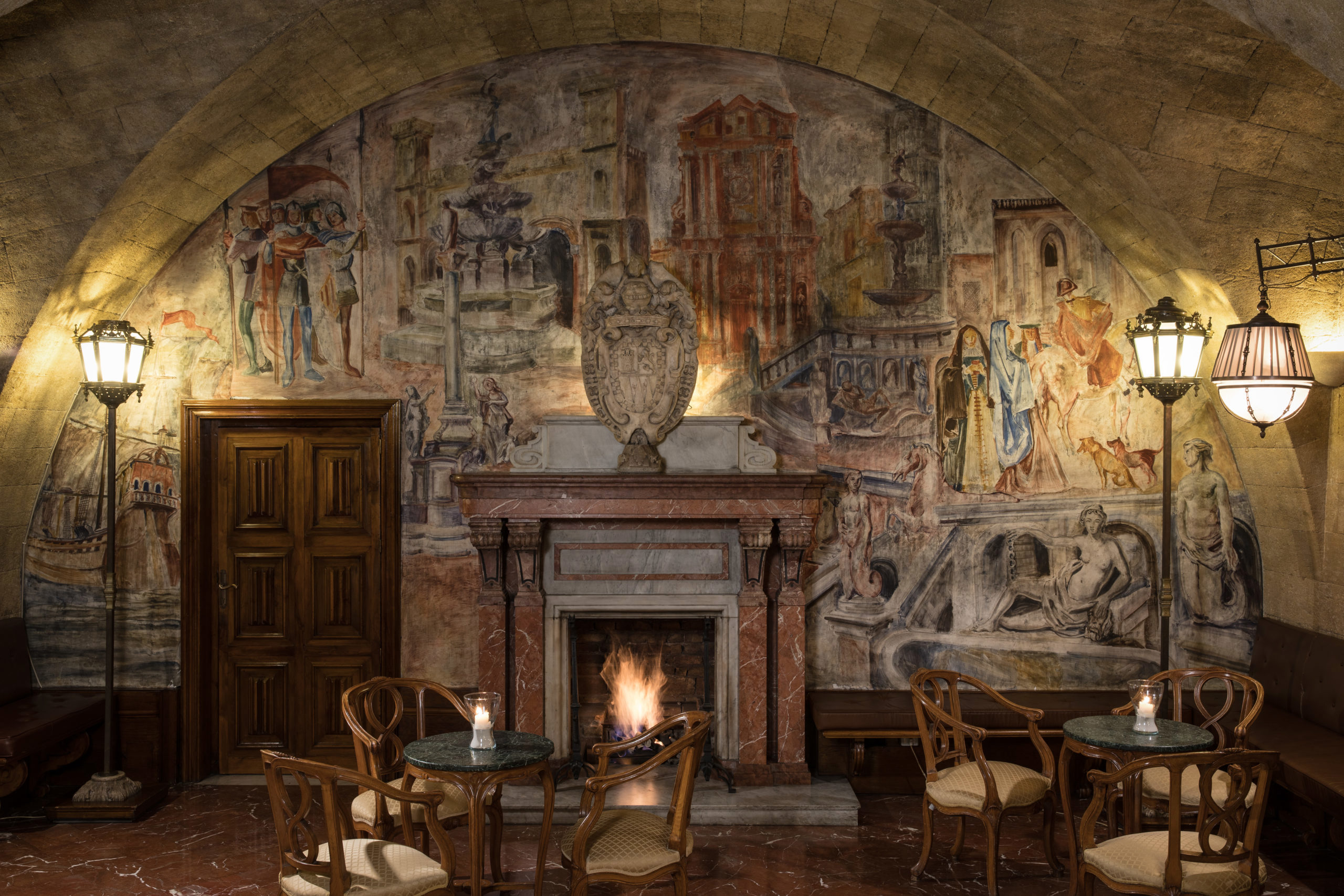 A moody room with a lit fireplace, furnished with wooden tables and chairs, under an arched ceiling adorned with a large, detailed mural depicting various figures and scenes.