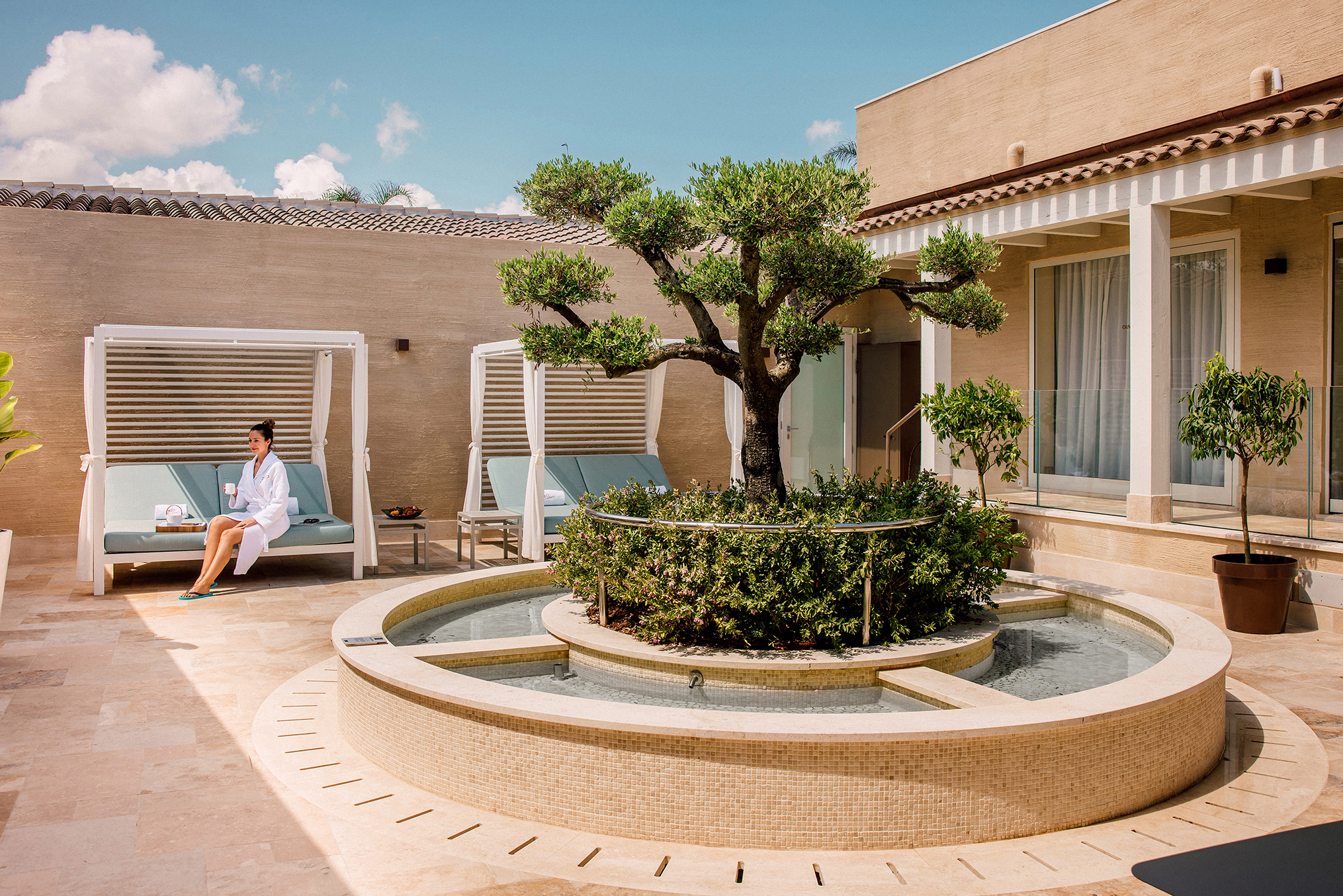 The courtyard of a spa with a central water feature and tree, with a woman sat under a shaded cabana