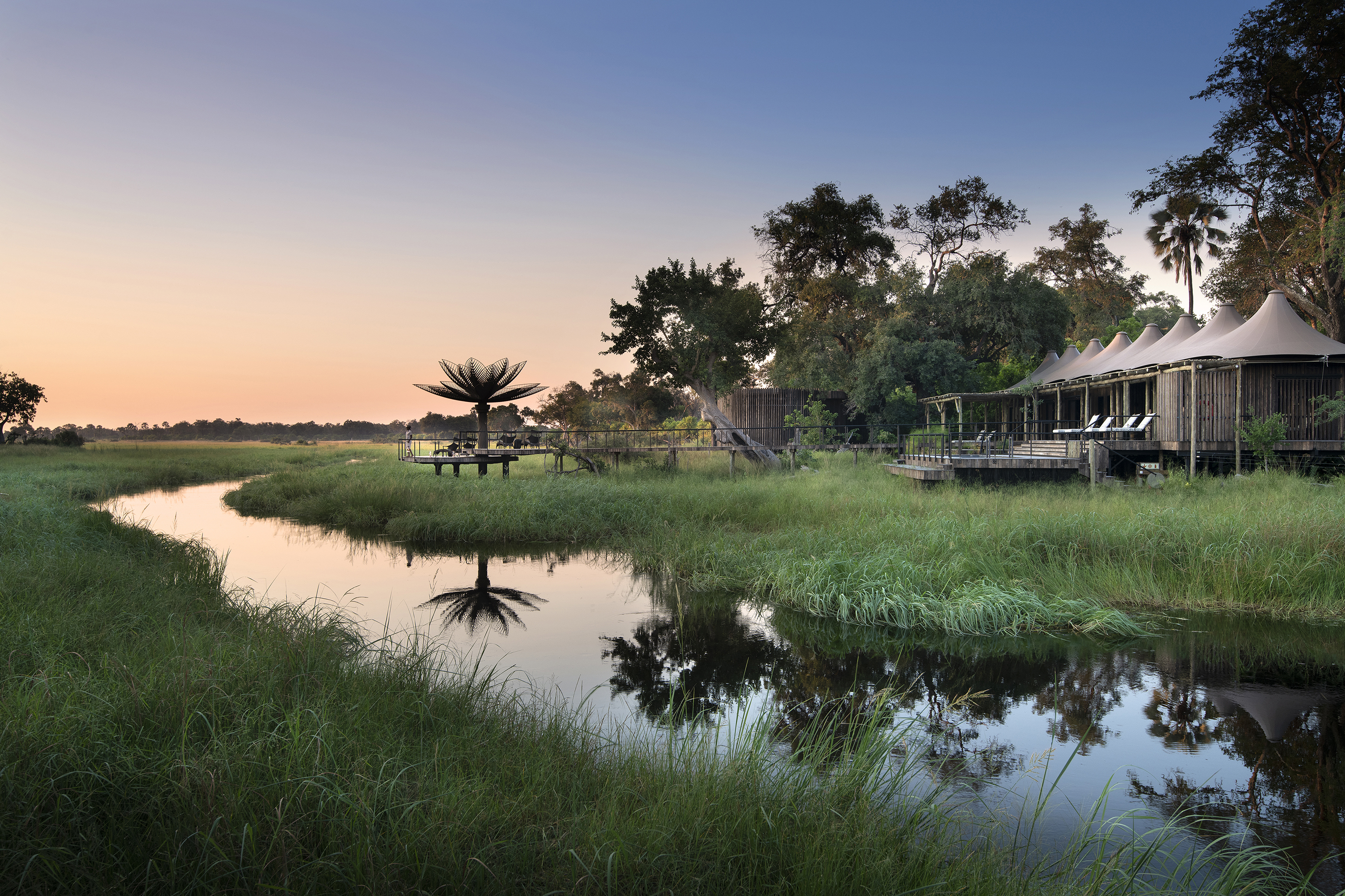 Tented exterior of Xigera Lodge facing a sweeping river surrounded by lush greenery at sunset with a viewing platform stretching out to the water