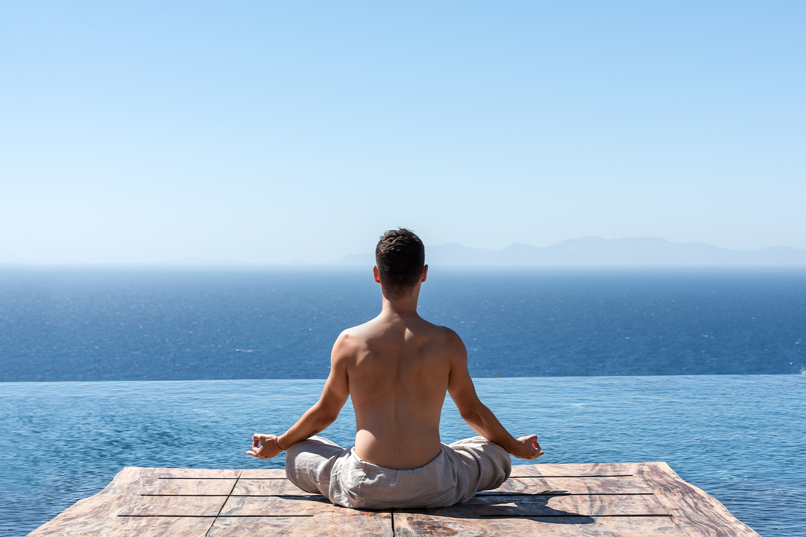 A man sits crossed legged, meditating with his back to the camera, on a wooden platform overlooking the ocean