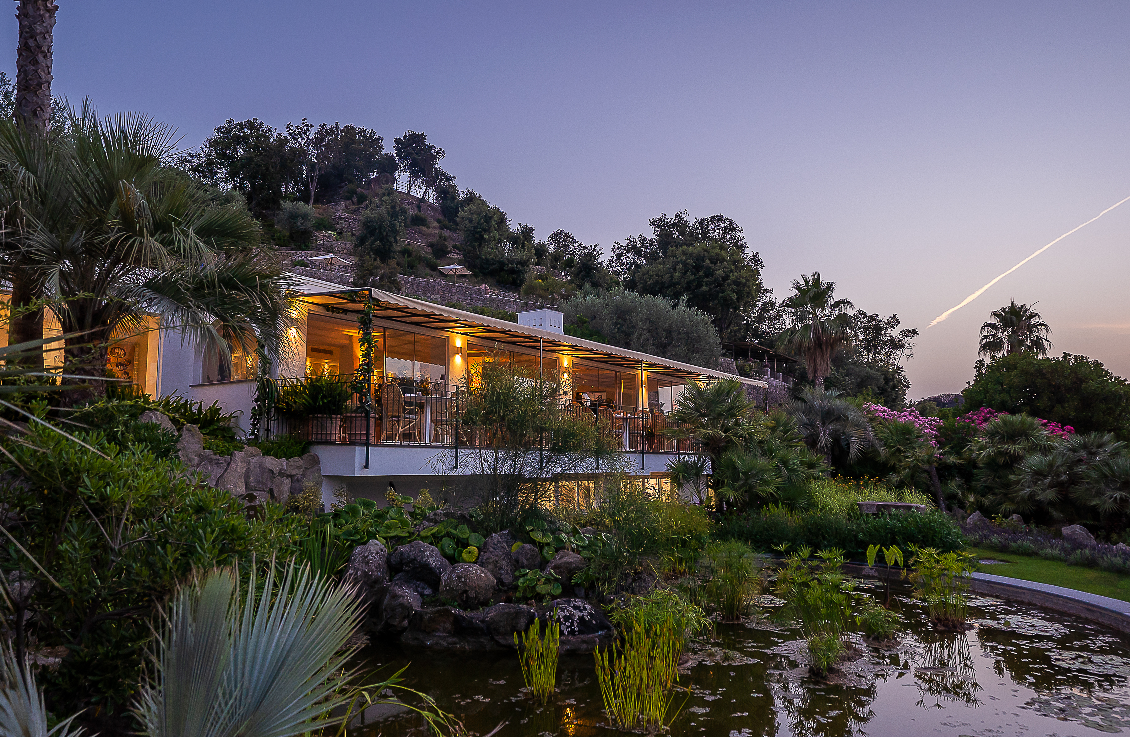 Mirto Fine Dining Restaurant at Botania Relais & Spa in Ischia at dusk, featuring a serene pond surrounded by lush greenery and a beautifully lit terrace.