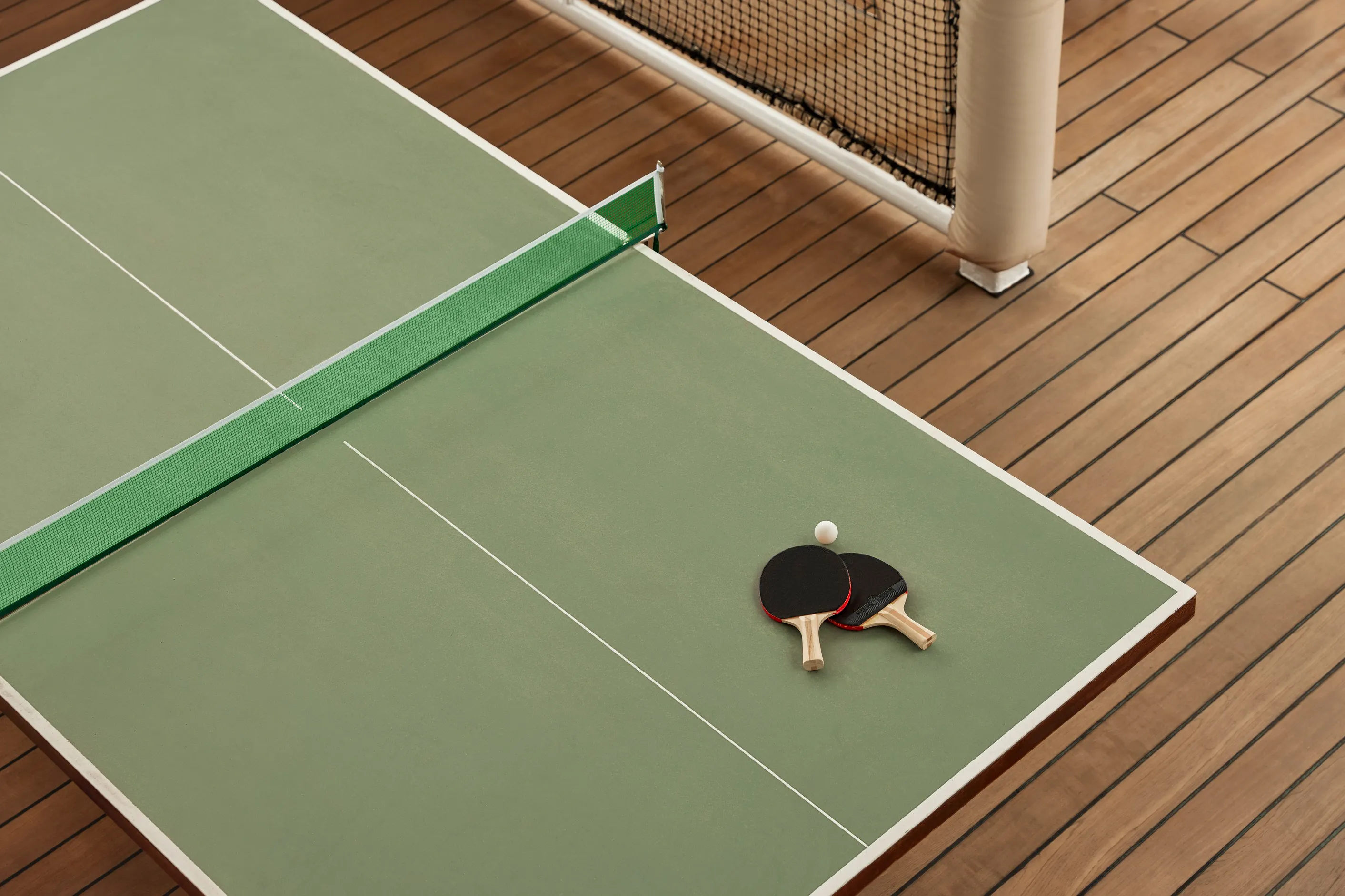 Two table tennis paddles and a ball resting on a green ping pong table with a wooden cruise deck beneath.