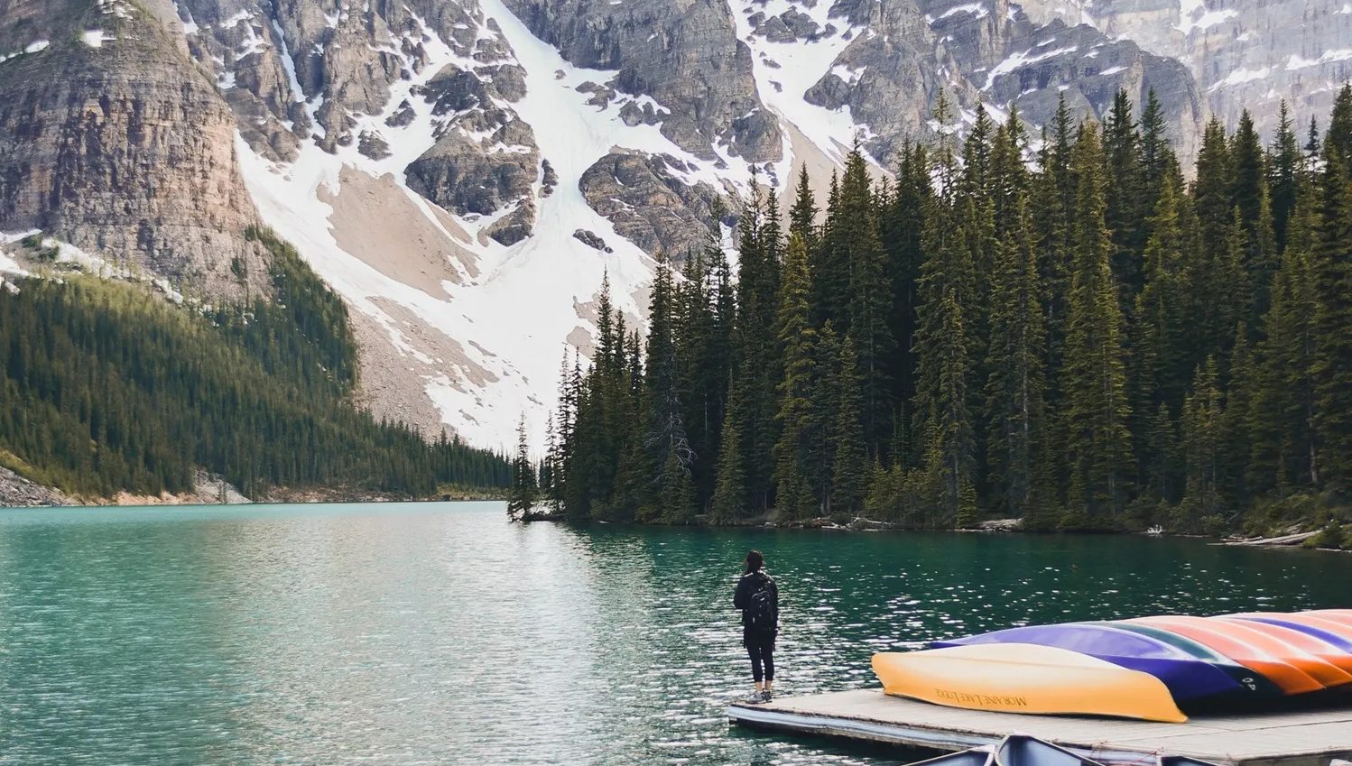 A person stands on a dock by a turquoise lake, surrounded by pine trees and snow-capped mountains.