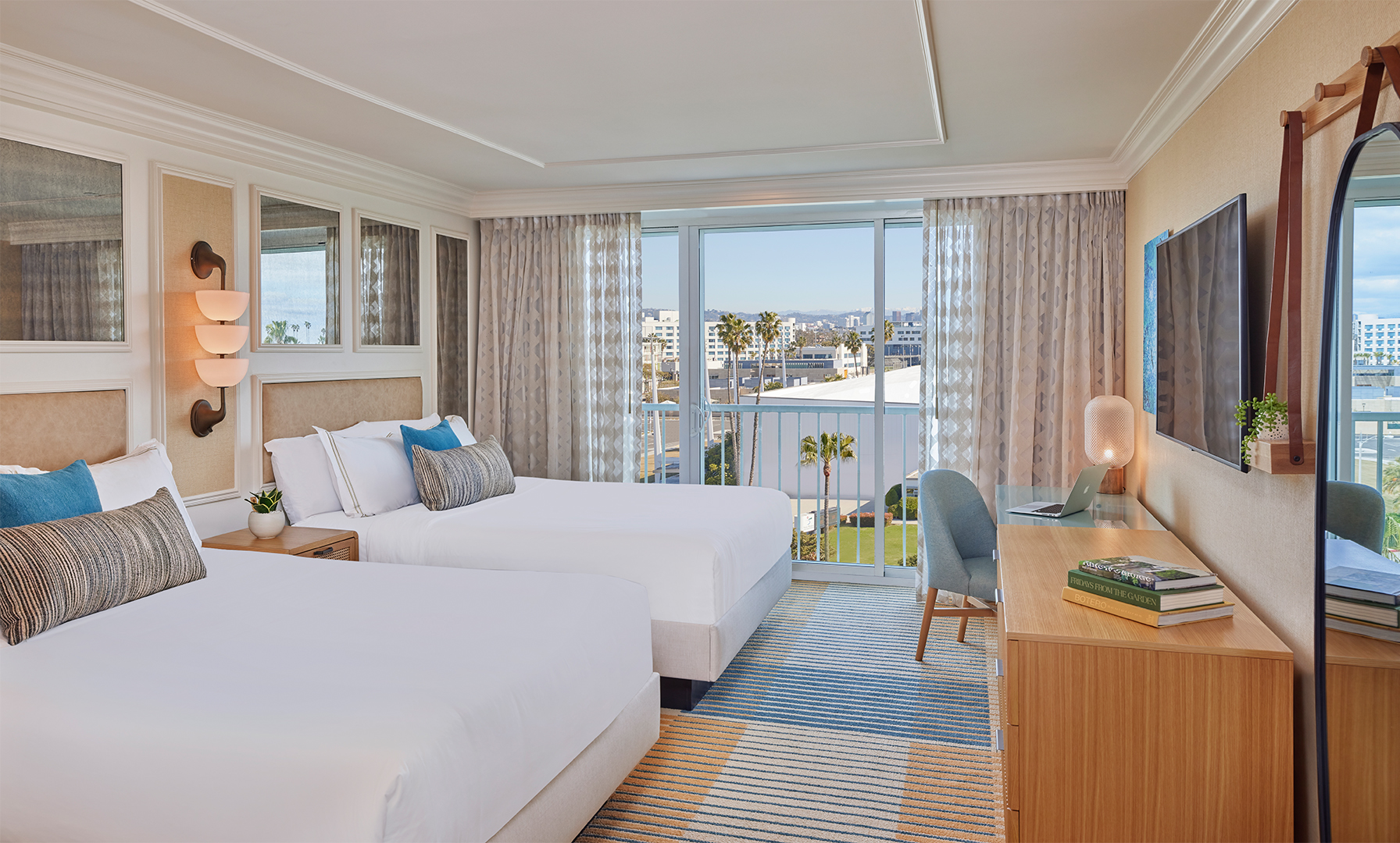 A twin hotel suite with a window view of a blue sky and ocean. The room is decorated in cream and brown tones and features twin double beds, a large chest of drawers, a TV and a blue arm chair.