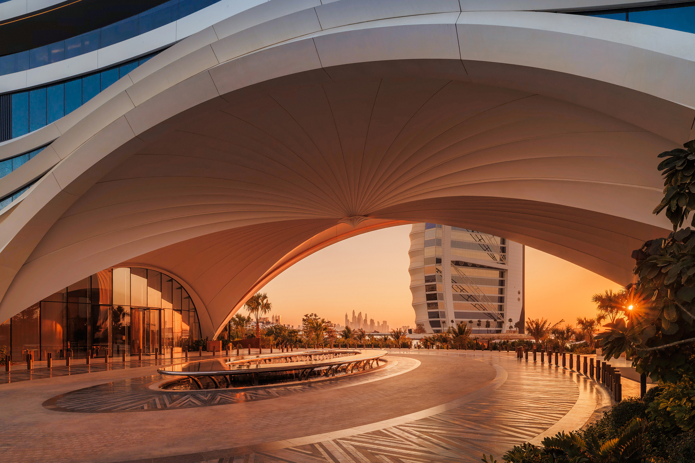 The futuristic arrival arch of Jumeirah Marsa Al Arab featuring a large fountain with a glimpse of the sunset and Burj Al Arab in the distance