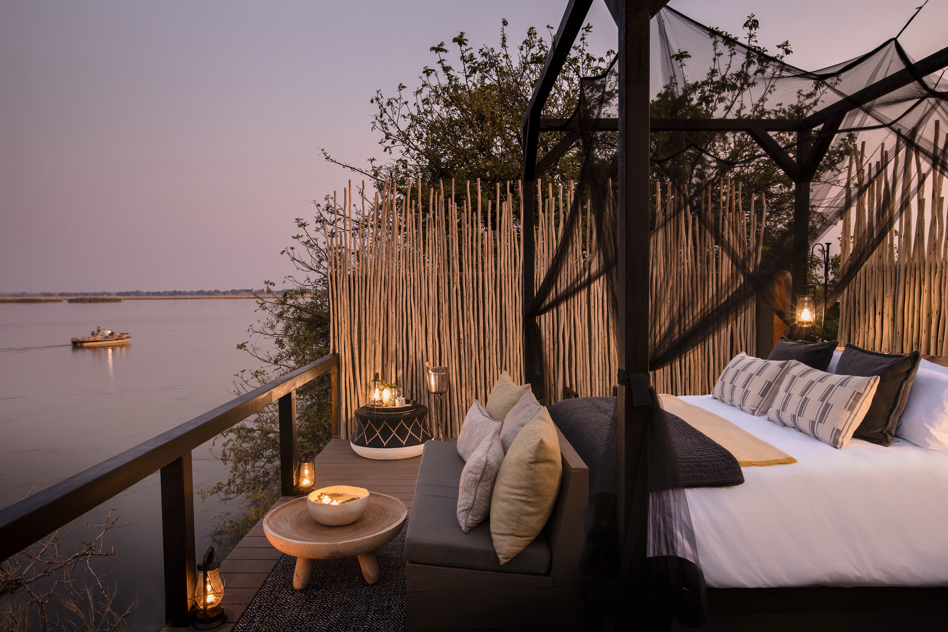 A large outdoor bed with dark netting and white sheets on a high deck with tables and candles overlooking the Zambezi River as a boat drifts past