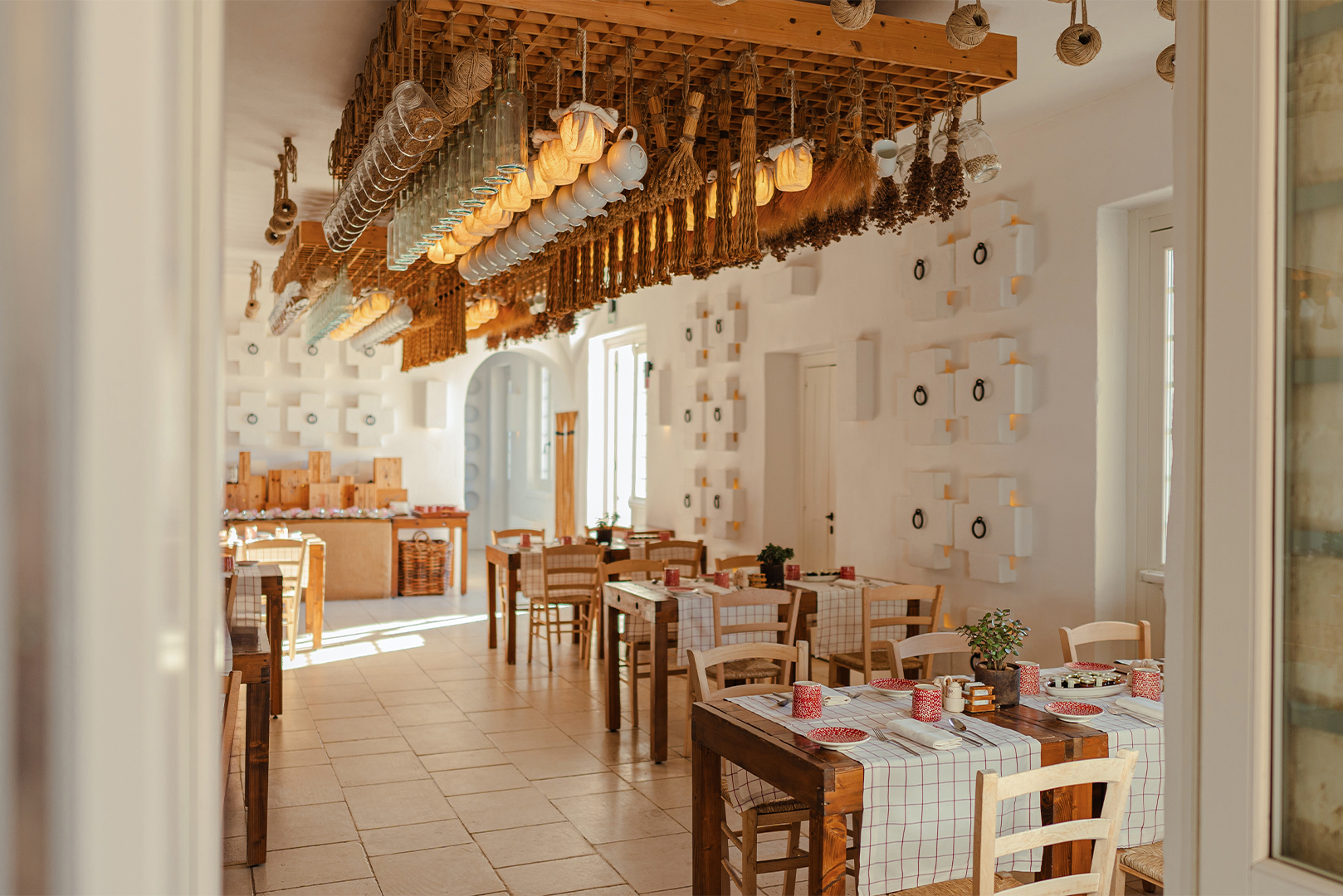 A bright and airy dining room with white walls decorated with small plates, wooden tables with checkered tablecloths, and a unique ceiling installation of hanging dried herbs and garlic.