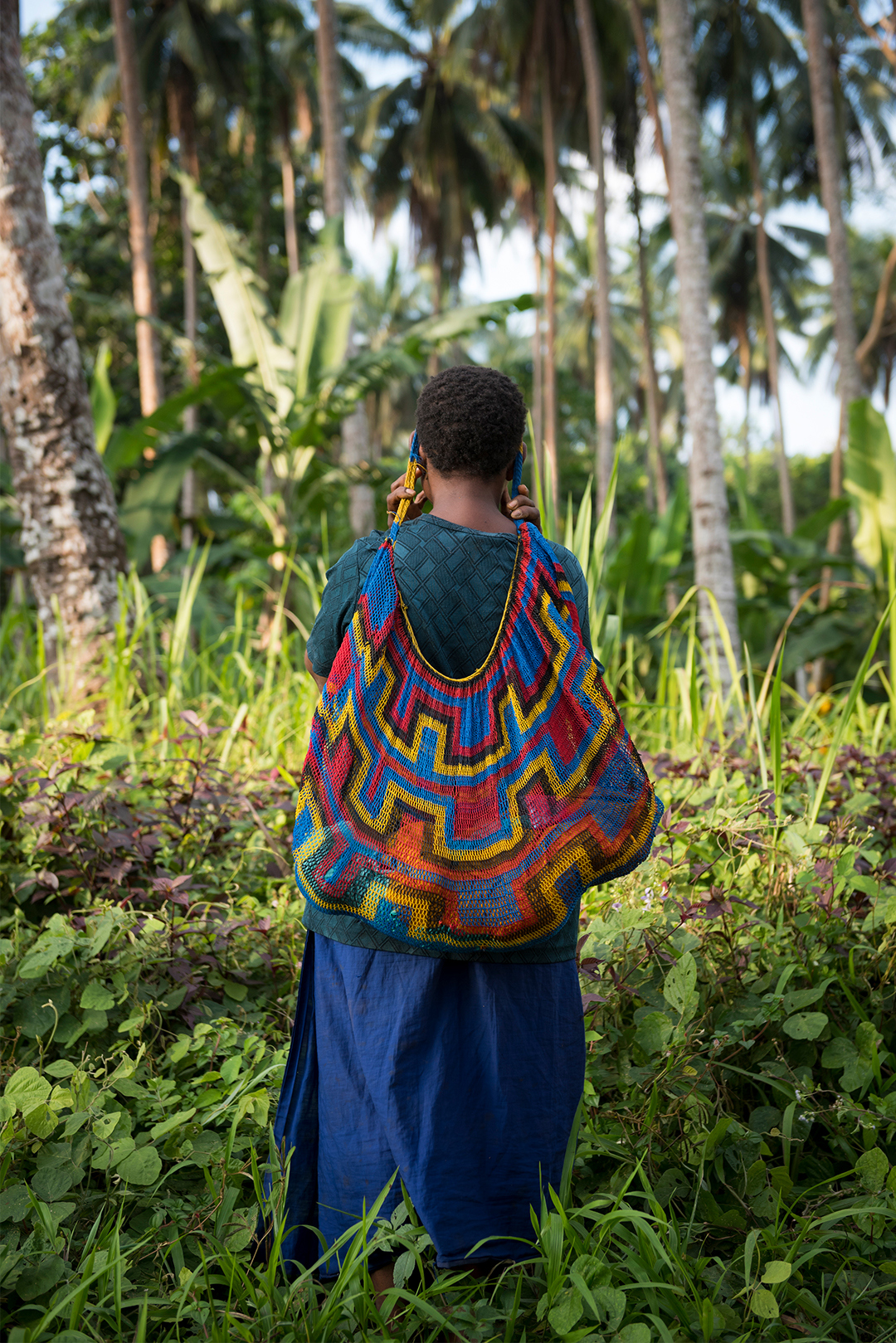 A person with a colourful patterned backpack stands amidst lush greenery, facing away from the camera.
