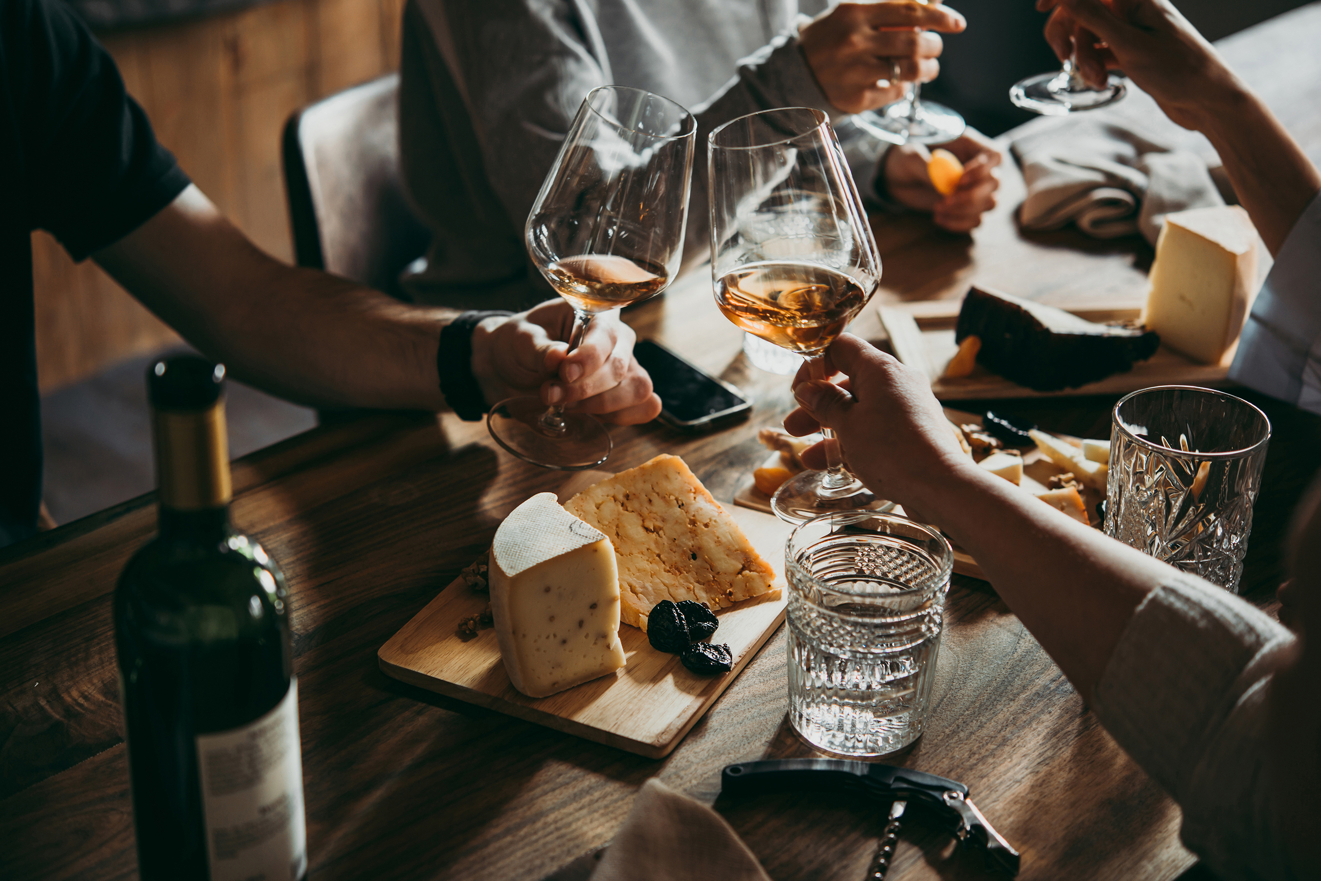 Four hands clinking rose wine glasses in cheers over a table set with cheese and ornate glasses of water