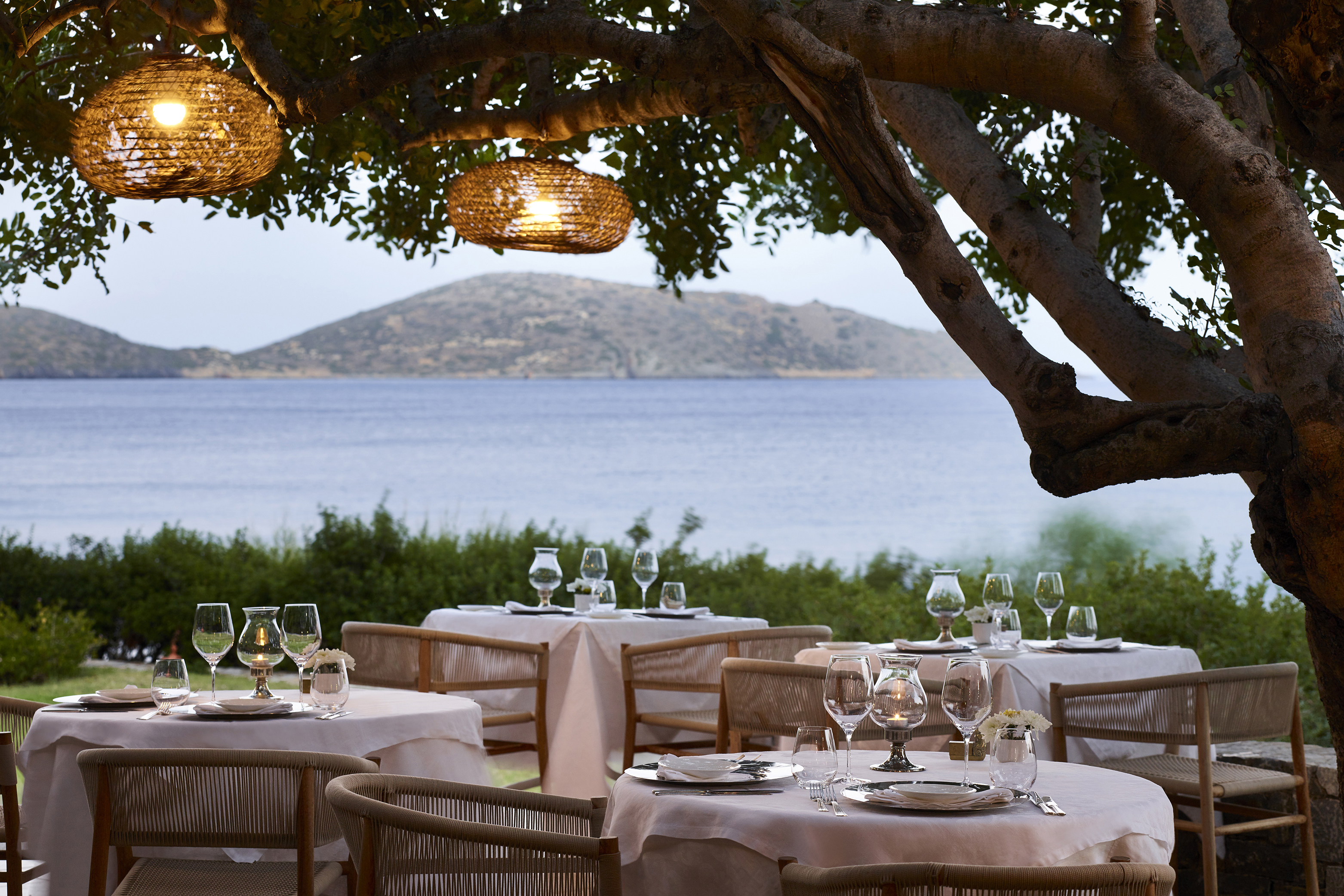 Four tables set for dinner below a tree with hanging lanterns looking out to sea
