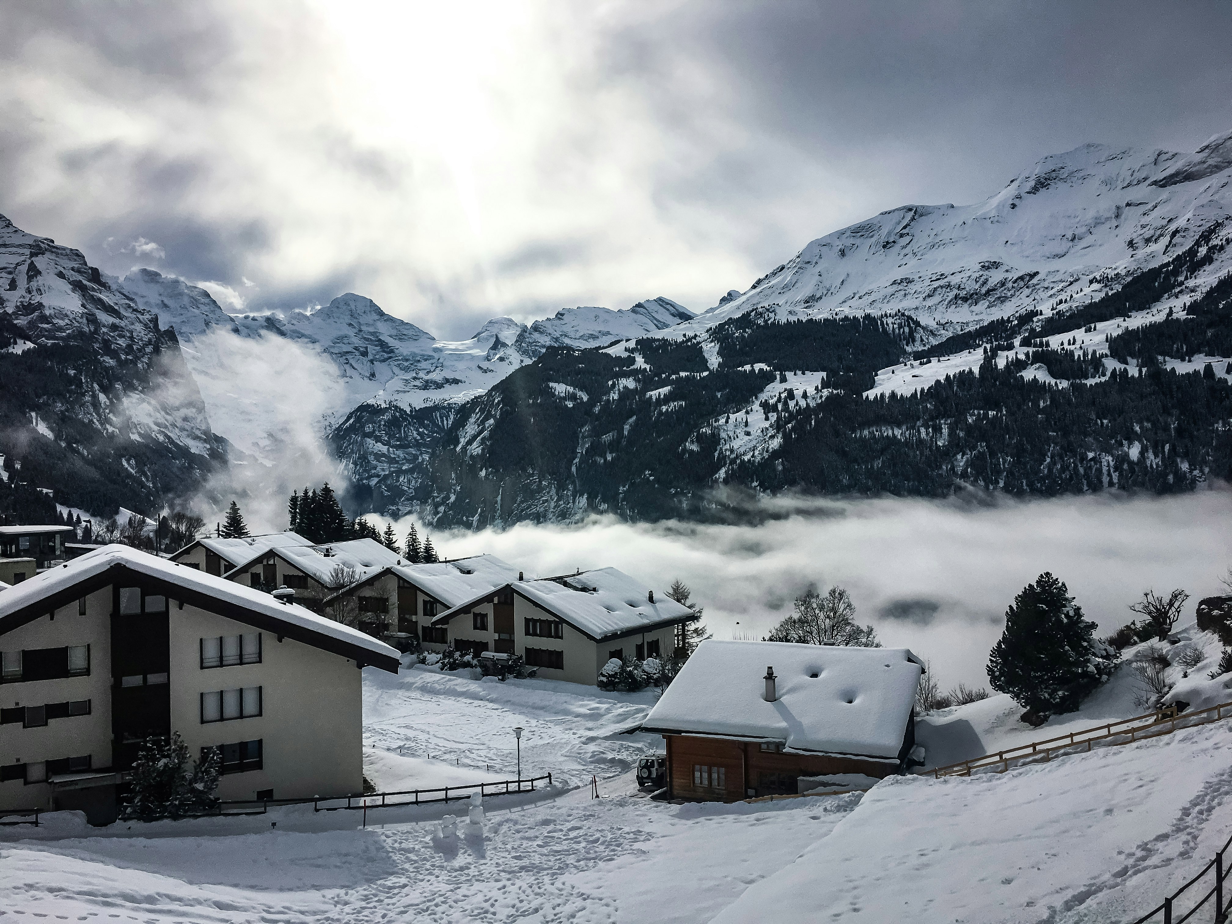 A snowy scene of Wengen in Switzerland with snow-covered buildings in front of white mountains with the sun breaking through the clouds