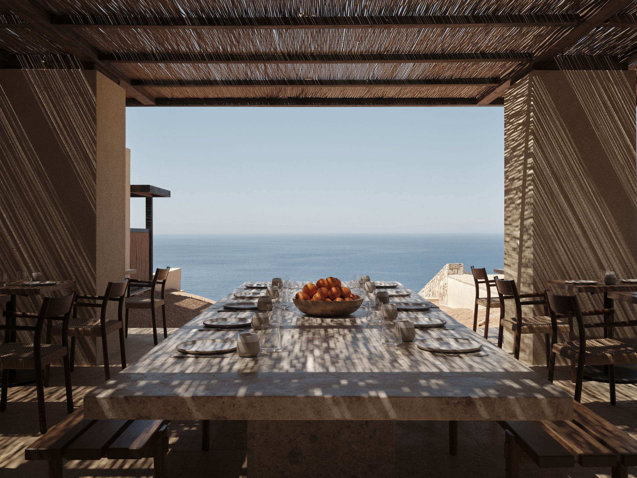 A long dining table speckled with shadows from the woven roof looking out to the open ocean