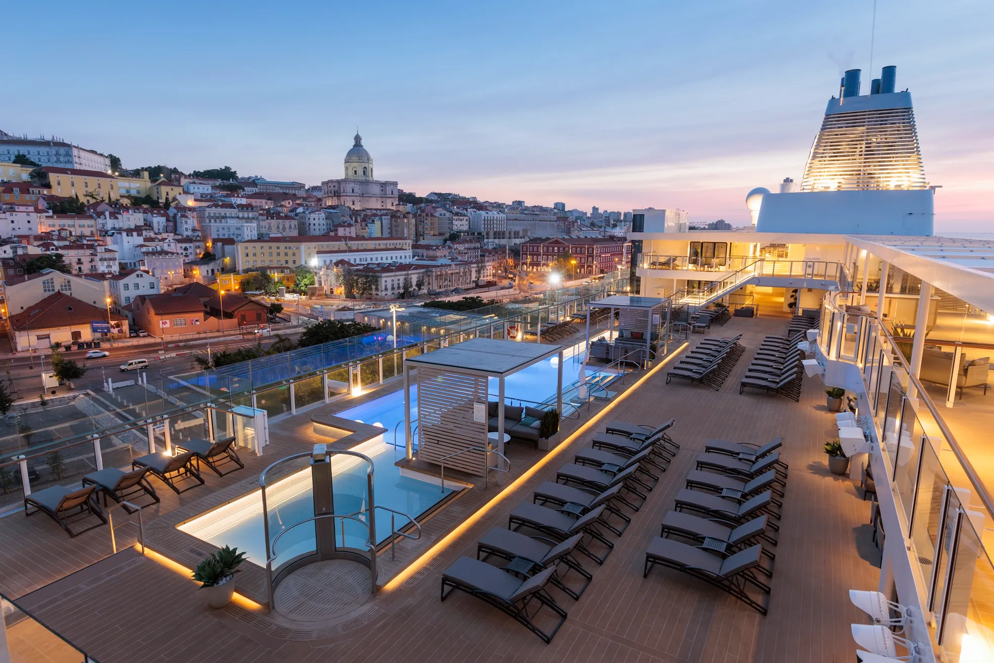 A top deck pool area with lounge chairs overlooking a cityscape at sunset.