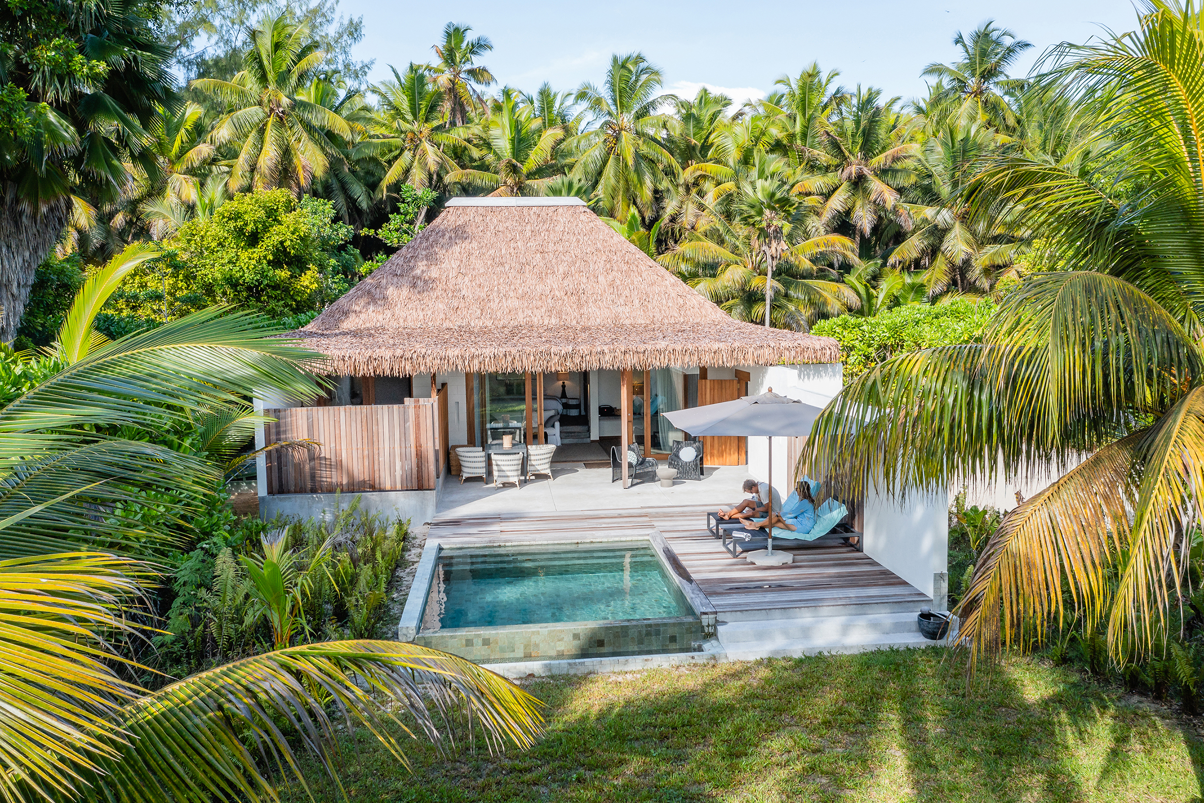 A stylish thatched villa with plunge pool and two people sat on loungers surrounded by greenery