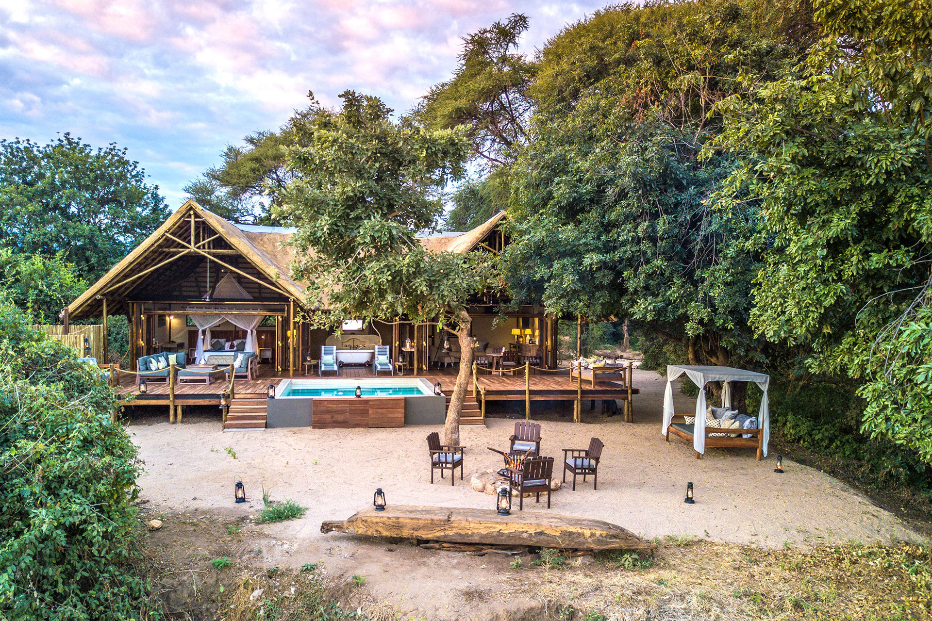 The outdoor area of a Safari Suite at Chiawa Camp featuring wooden chairs around a firepit and cabana before a thatched building with a pool on the deck