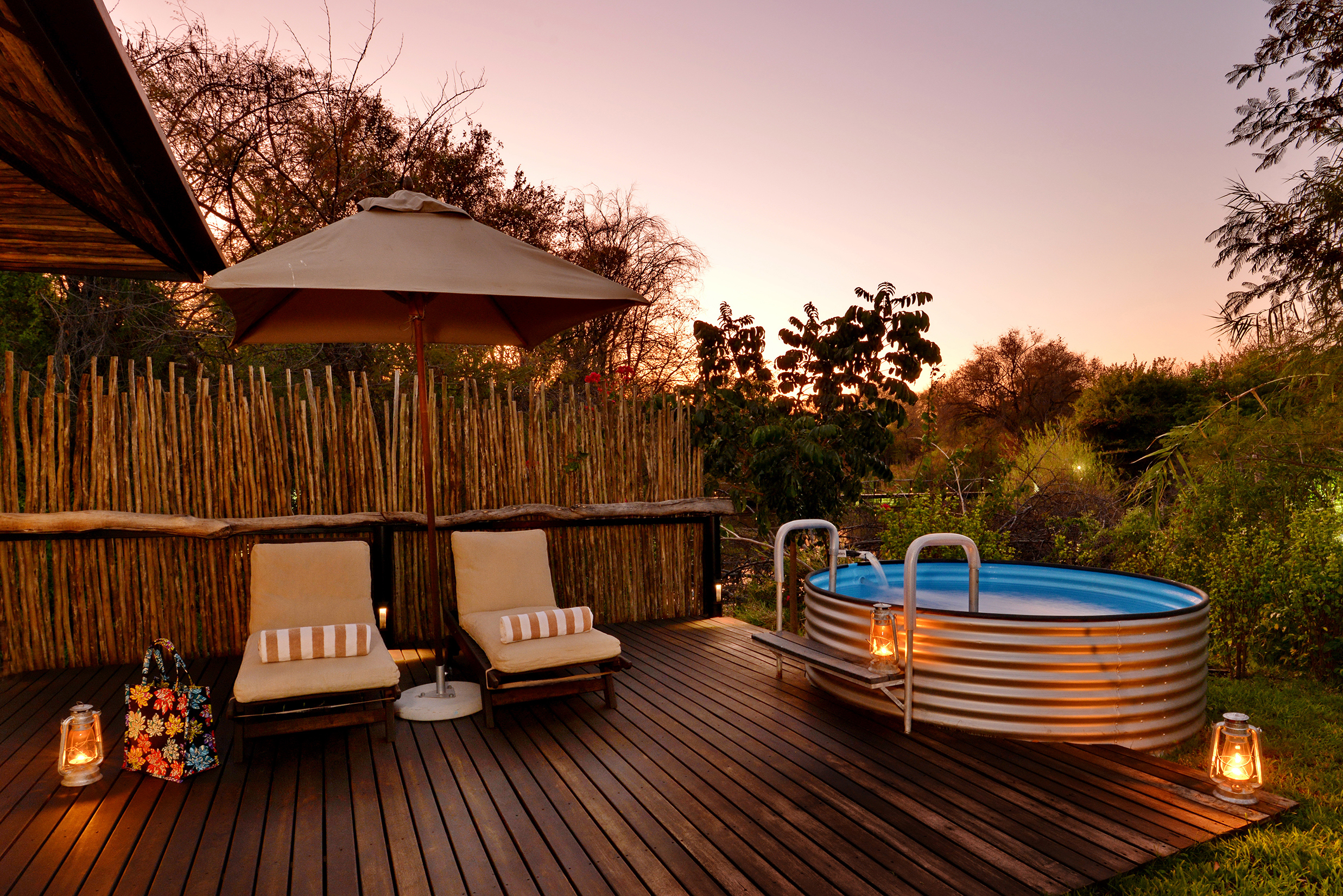 A serene outdoor deck at Mukwa River Lodge with cushioned lounge chairs, a circular metal plunge pool, lanterns, and lush greenery at sunset.