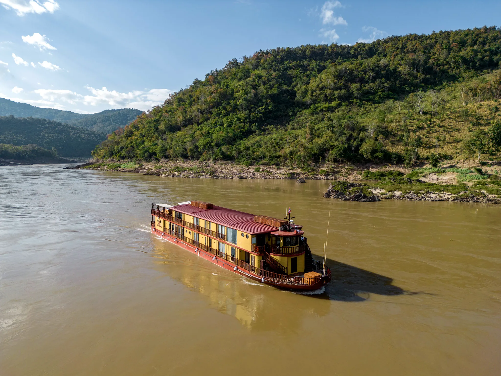A vibrant Heritage Line riverboat cruises along a wide, muddy river with lush green hills in the background under a partly cloudy sky.