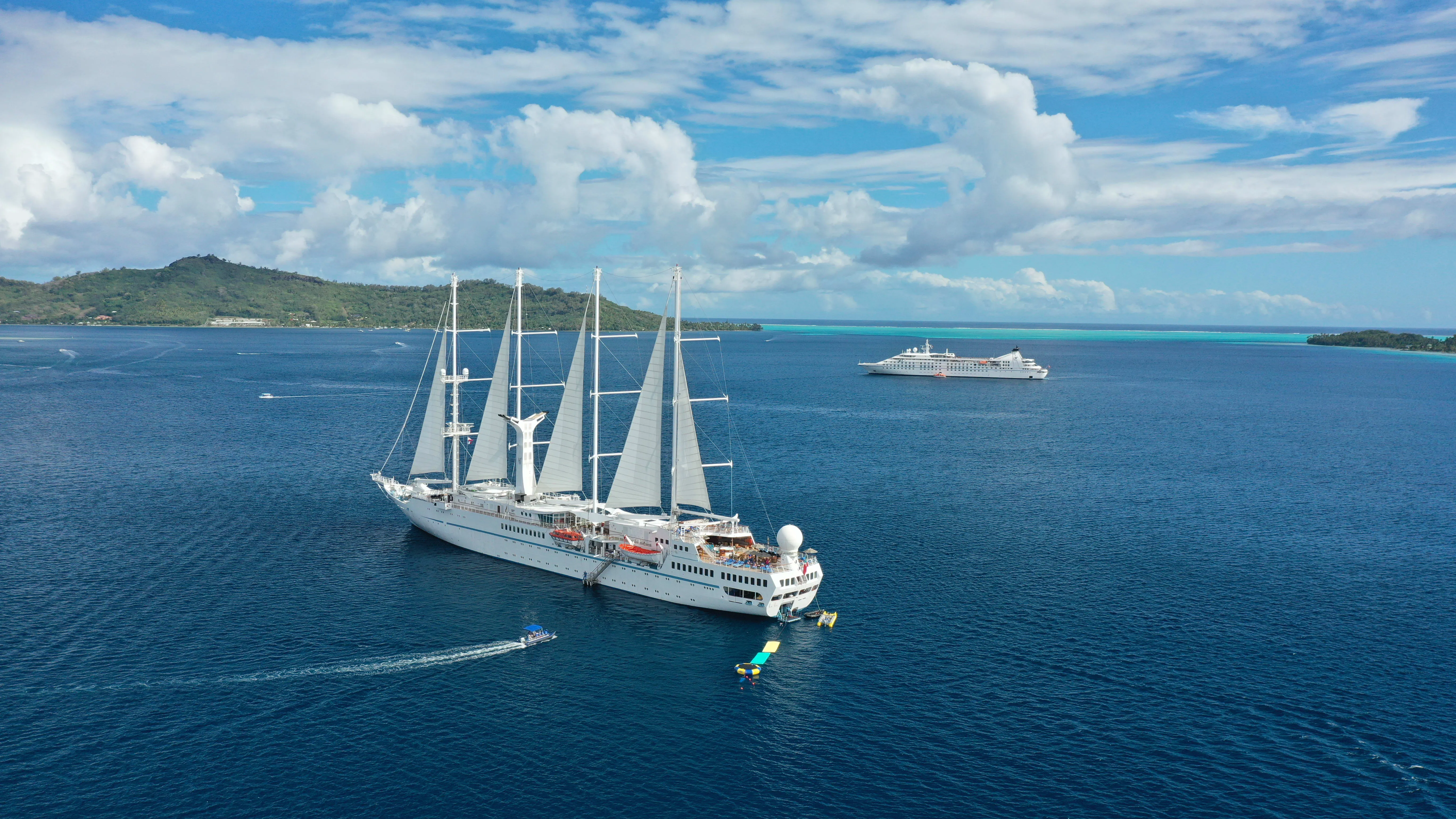 A large white sailing ship anchored in calm blue sea near a smaller boat, with another cruise ship and a lush island in the distance.