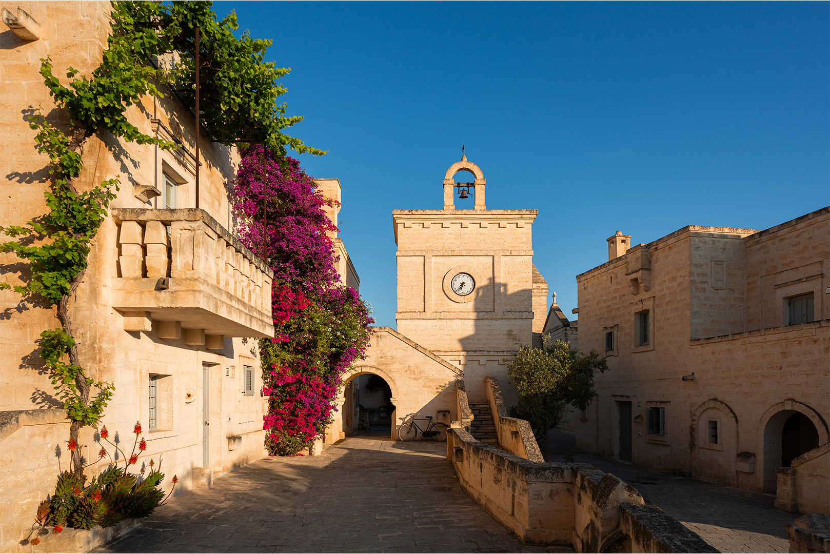 A stone pathway leads through an old town with traditional limestone buildings, vibrant bougainvillea, and a clock tower under a clear blue sky.