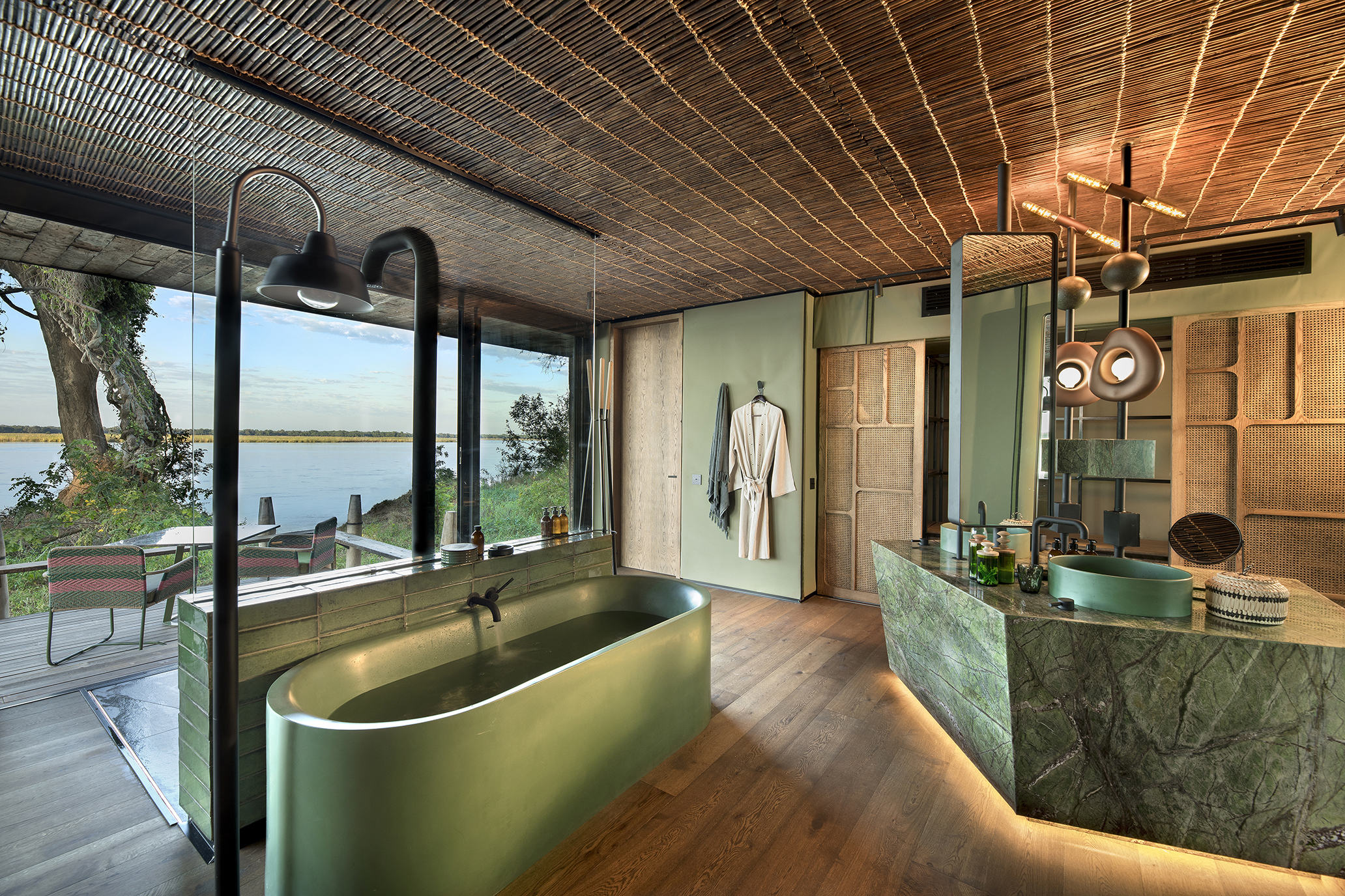 The bathroom of a suite at Lolebezi featuring green marble and stone beneath a thatched roof with a glimpse of the river beyond