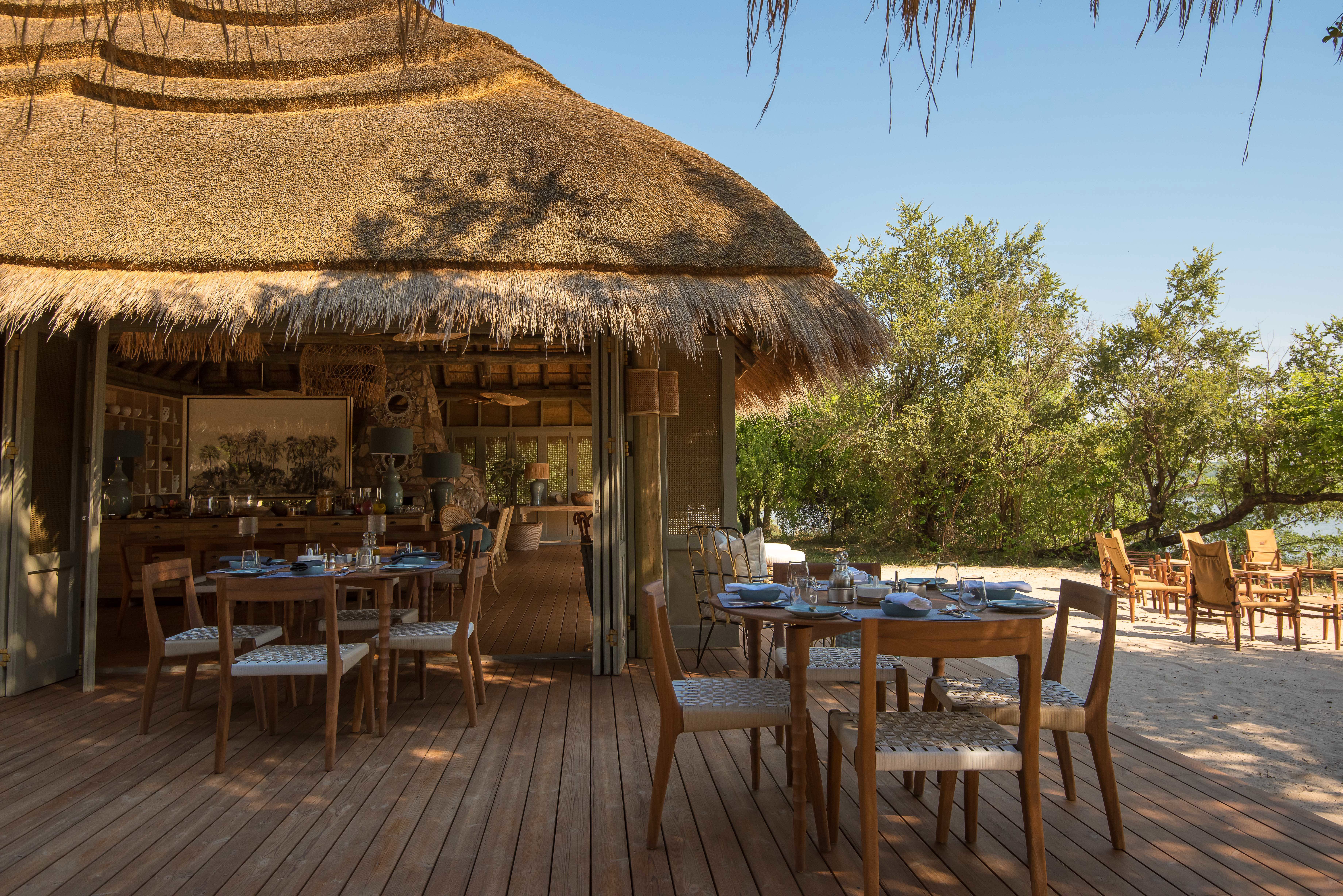 A dining area beside a sandy riverbank featuring outdoor tables on a deck and a thatched open-sided building with tables inside