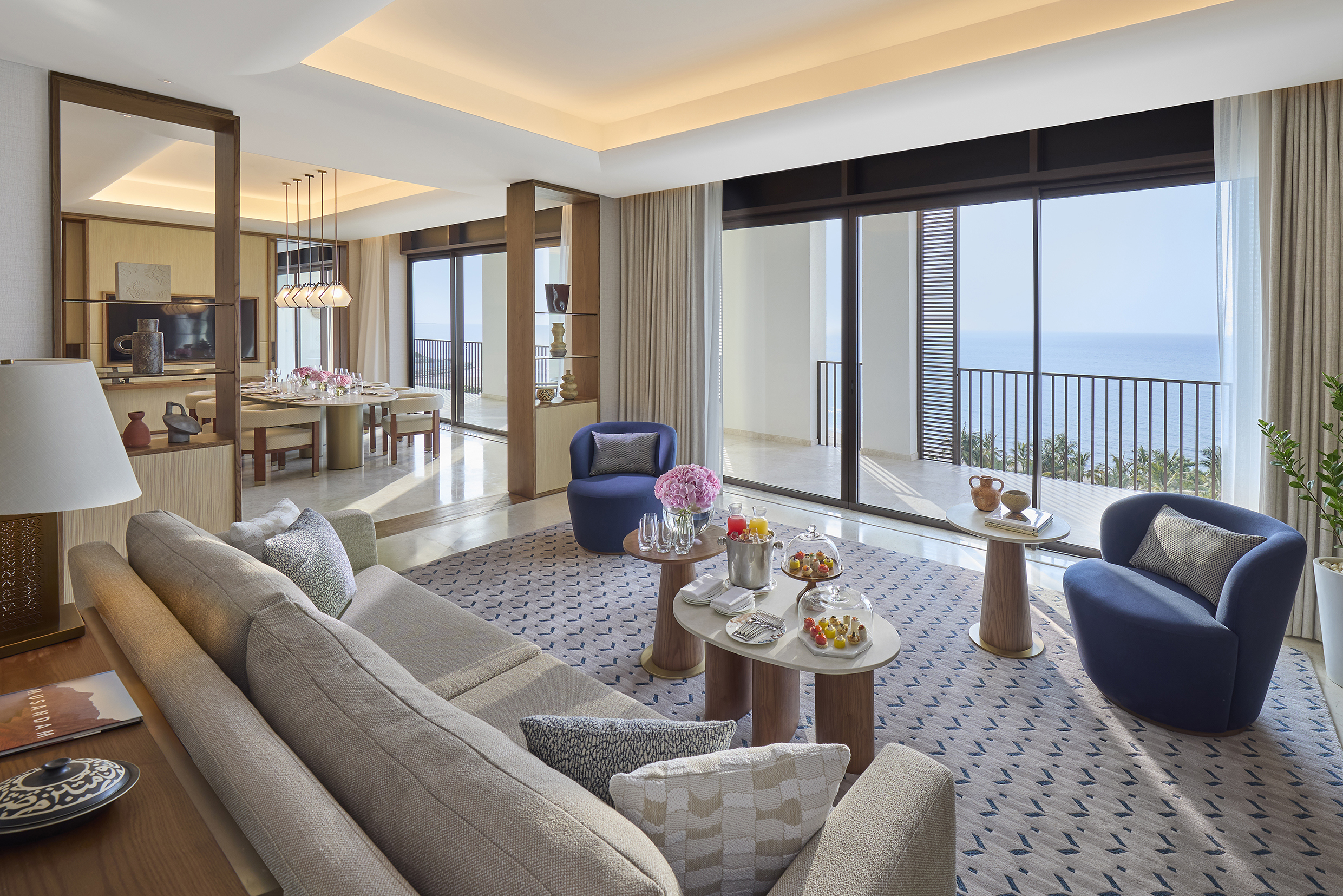 The large lounge of a suite featuring big windows and comfy furniture, with a glimpse of a dining room behind