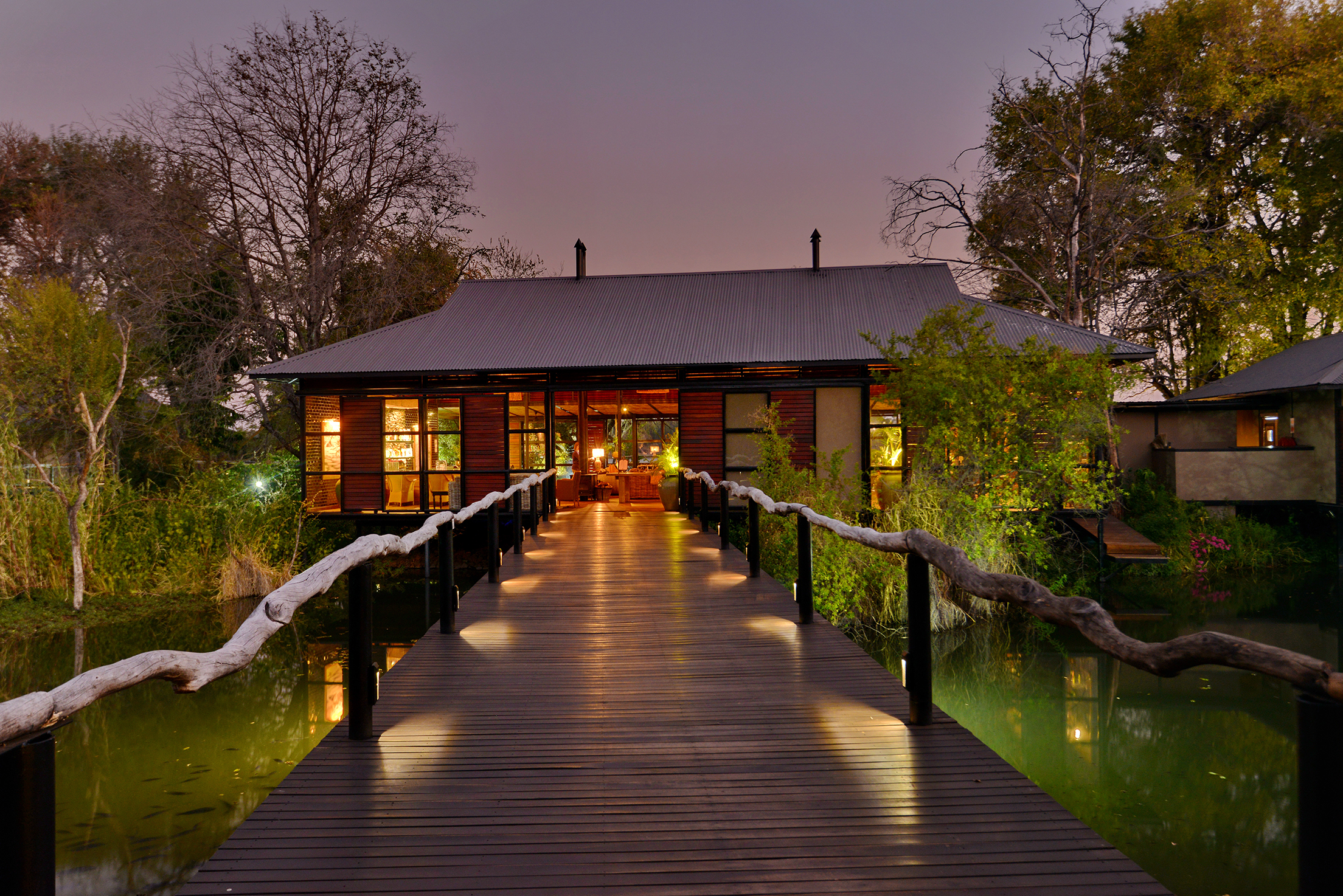 The exterior of Mukwa River Lodge across wooden walkway with rustic railings leads to a warmly lit lodge surrounded by trees and water at dusk