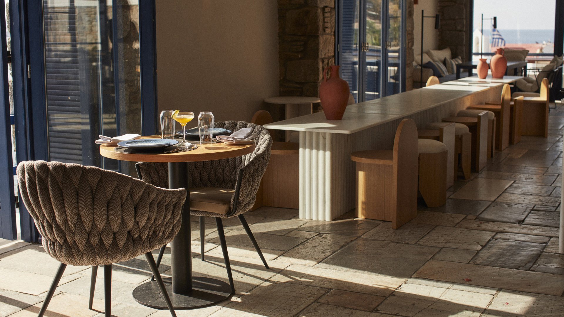 A stylish table for two set for breakfast bathed in sunlight from a nearby window, sitting before a long communal table with stools