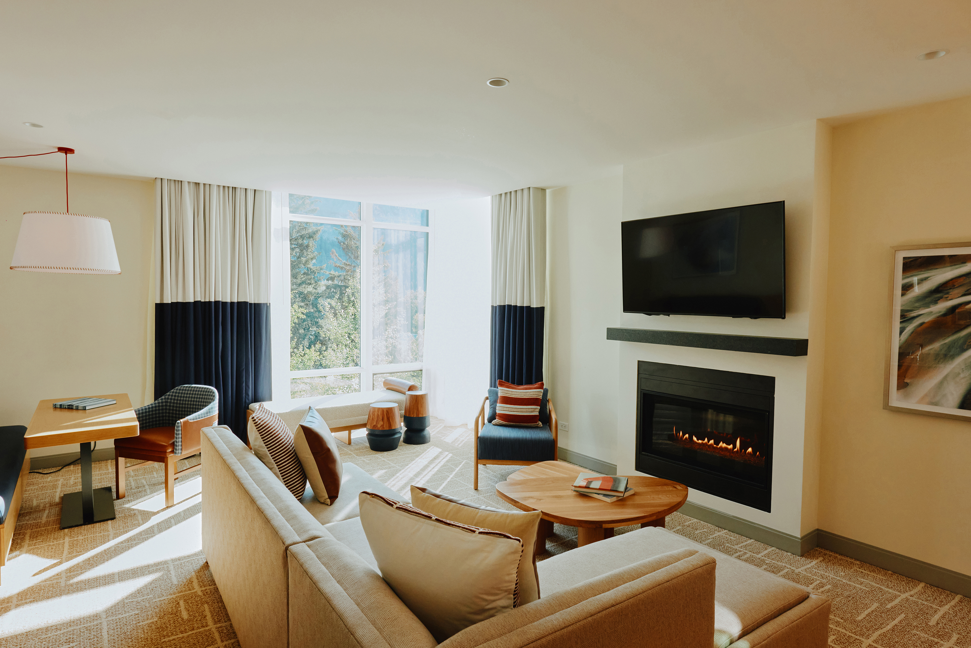 A modern hotel suite living room at Limelight Ketchum featuring a beige sofa, wooden coffee table, fireplace with a wall-mounted TV, and large windows with curtains