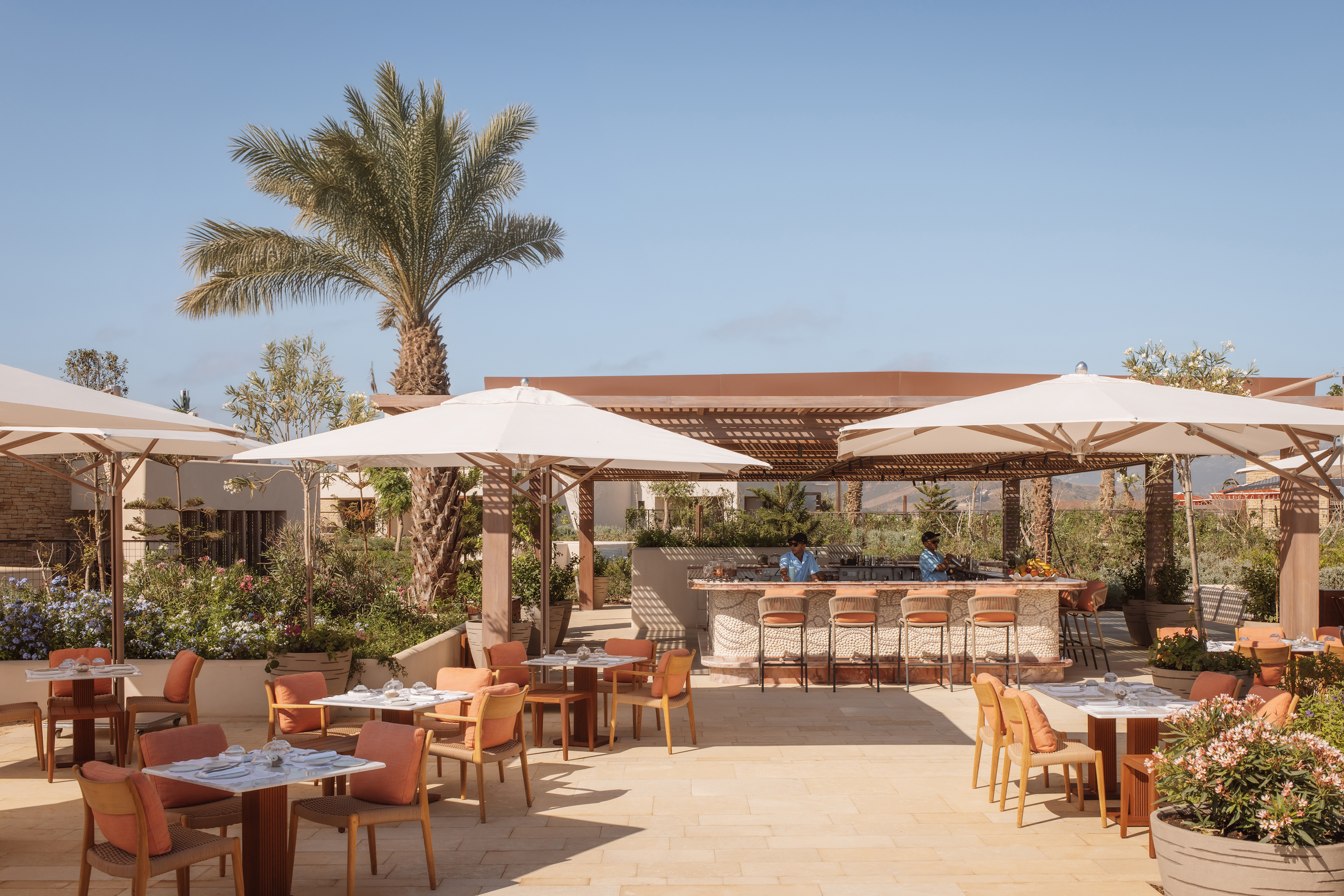 The outdoor Pool Beach bar at Royal Mansour Tamuda Bay featuring chic wooden dining tables with umbrellas, set up in front of a shaded bar with palm trees behind