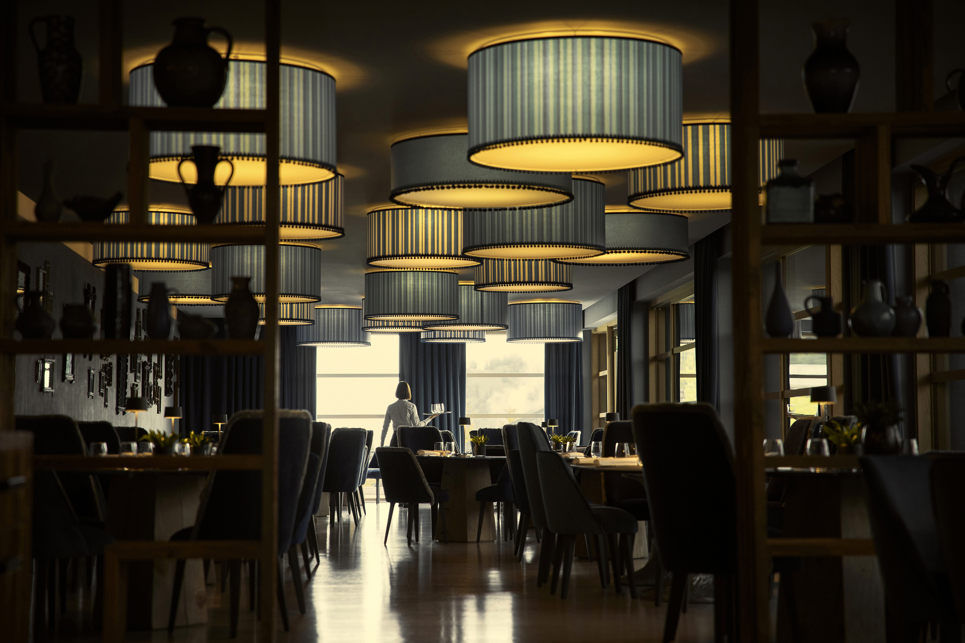 Sassolungo restaurant's elegant interior with round hanging lamps, set tables, and a silhouette of a person standing by the window.