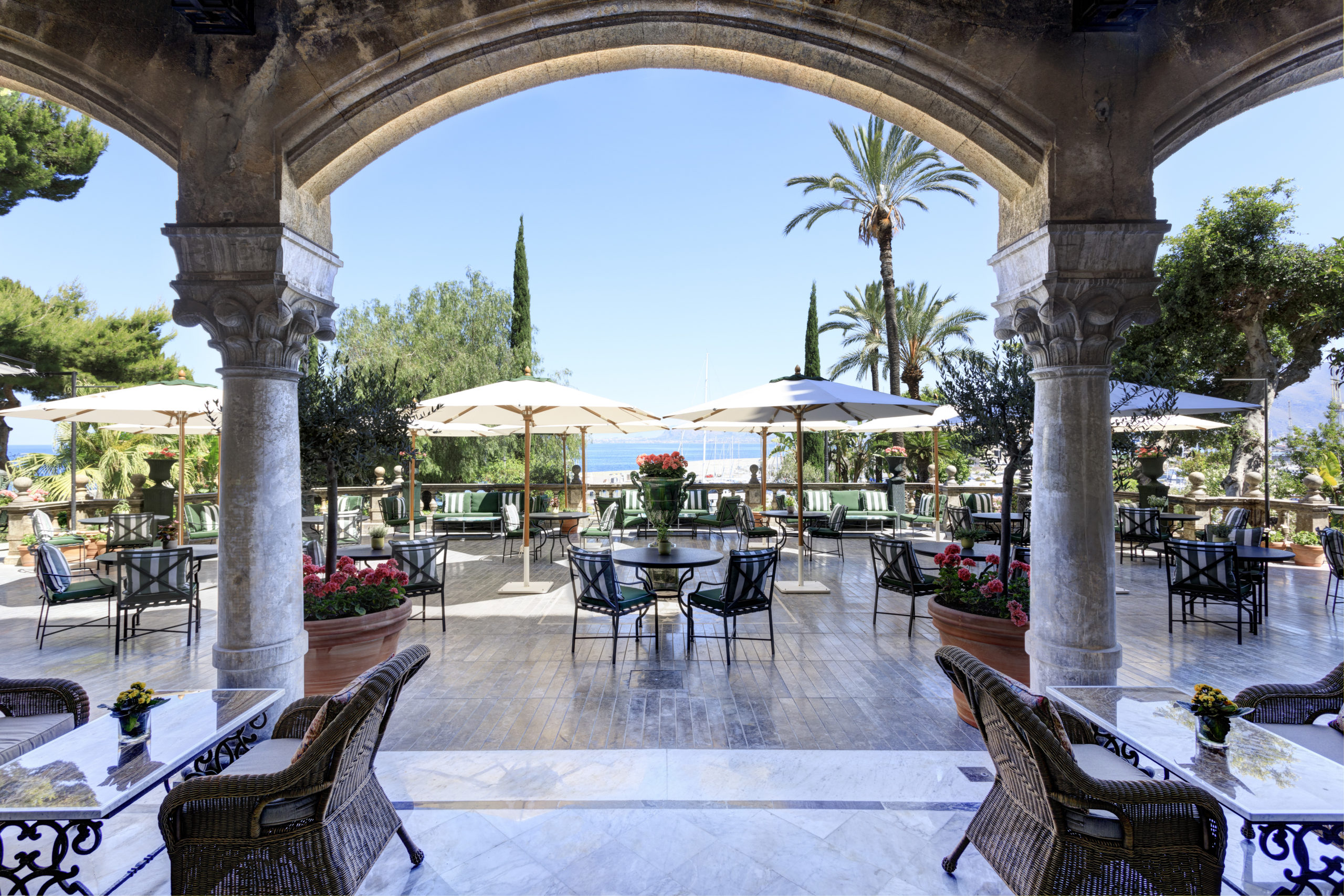 An ornate outdoor bar terrace with arched stone columns, wicker chairs, and white umbrellas set against a backdrop of lush greenery and clear blue sky.