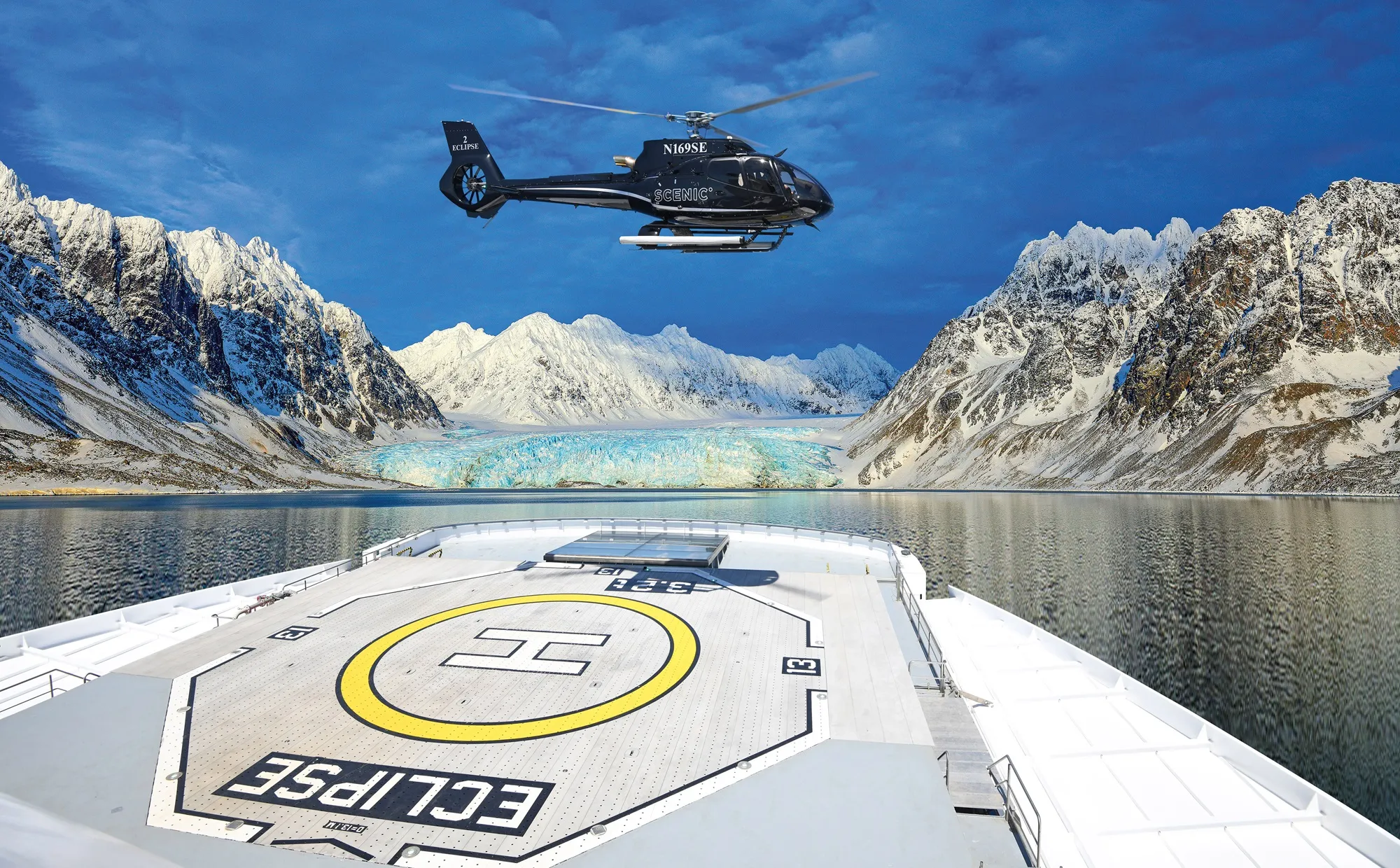 Helicopter hovering above the Scenic Eclipse ship’s helipad with snowy mountains and a glacier in the background.