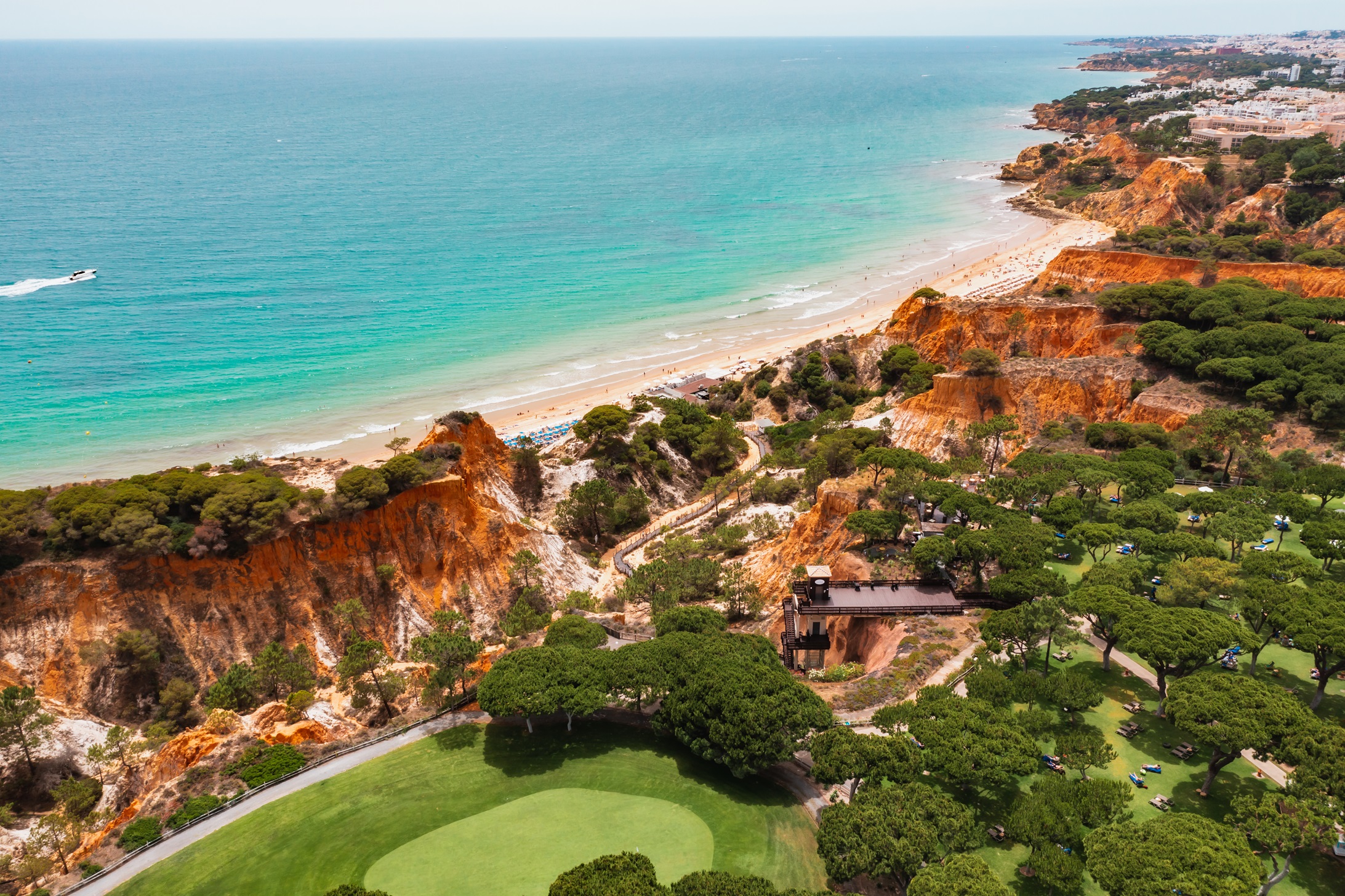 A coastal landscape featuring a golden sandy beach, turquoise water, reddish-brown cliffs with greenery, a golf course in the foreground, and buildings along the distant shoreline