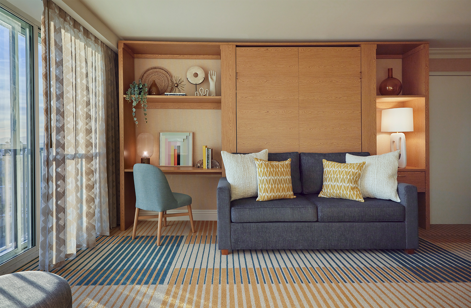 A living room of a hotel suite. The room is decorated in cream and blue tones with light wooden furniture. It features a desk and sofa.