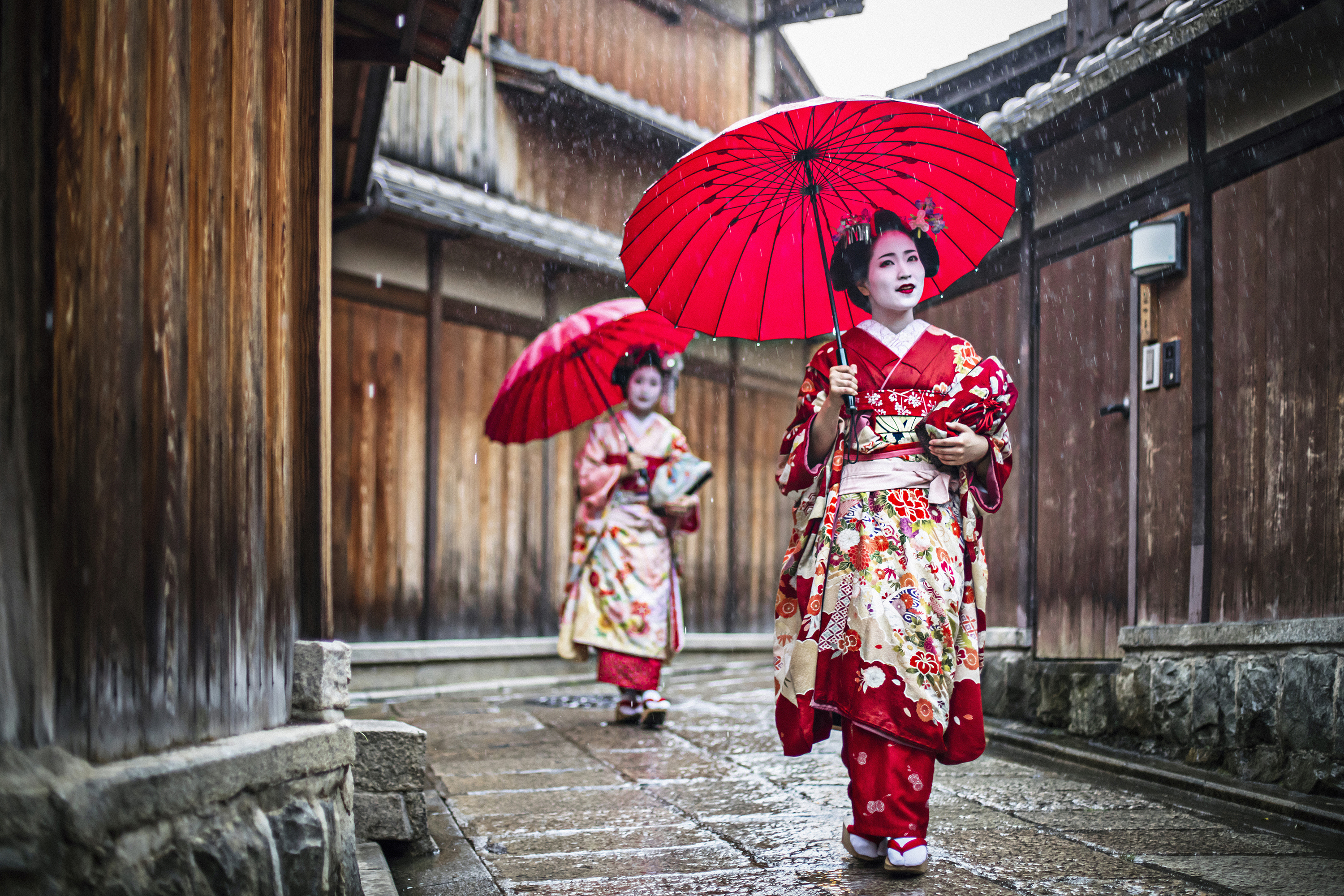 Two women in traditional geisha attire and makeup walking through the streets of Kyoto with red umbrellas