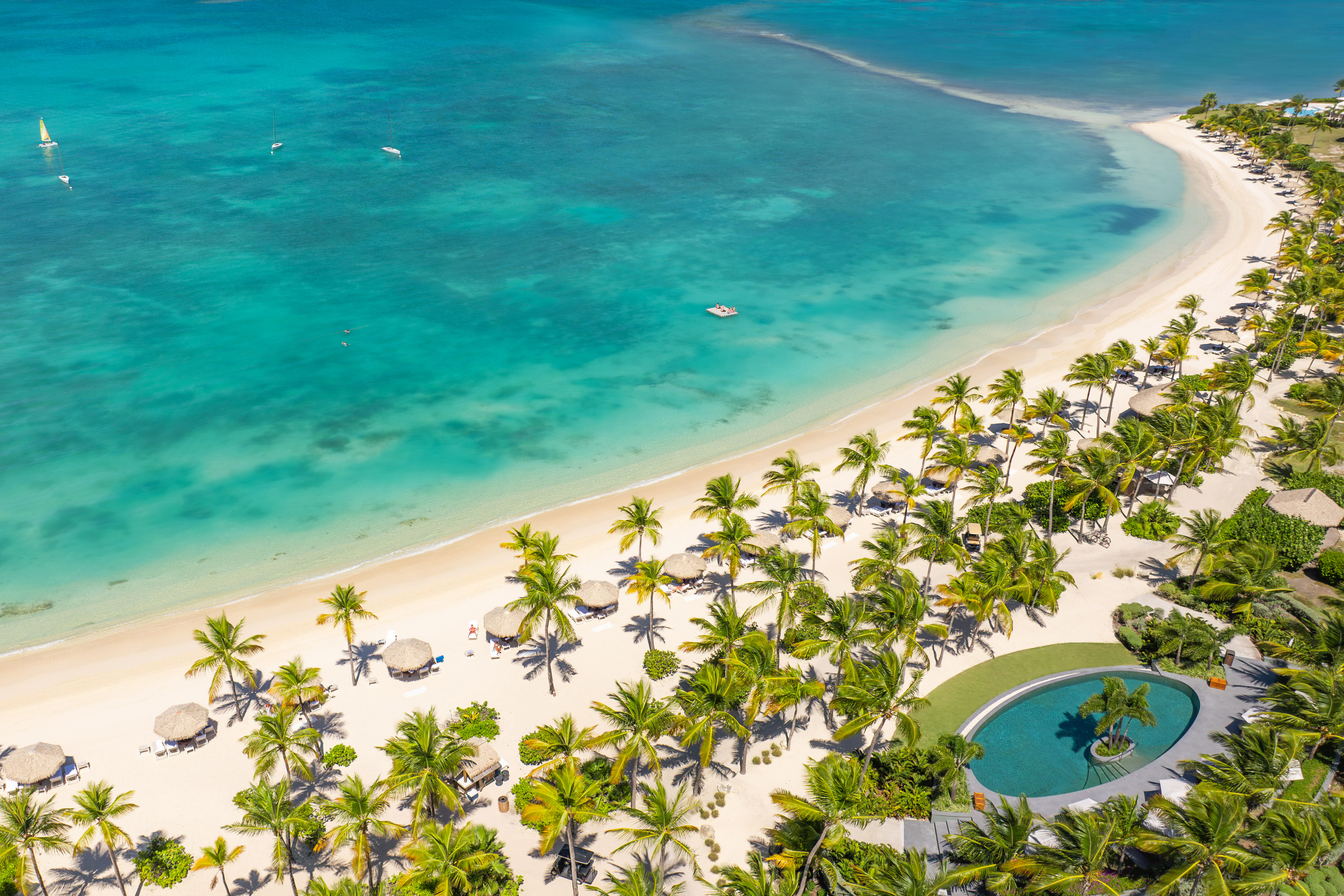 A birds eye view of a large, curved white sand beach on Jumby Bay Island flanked by the sea, palms and an oval swimming pool
