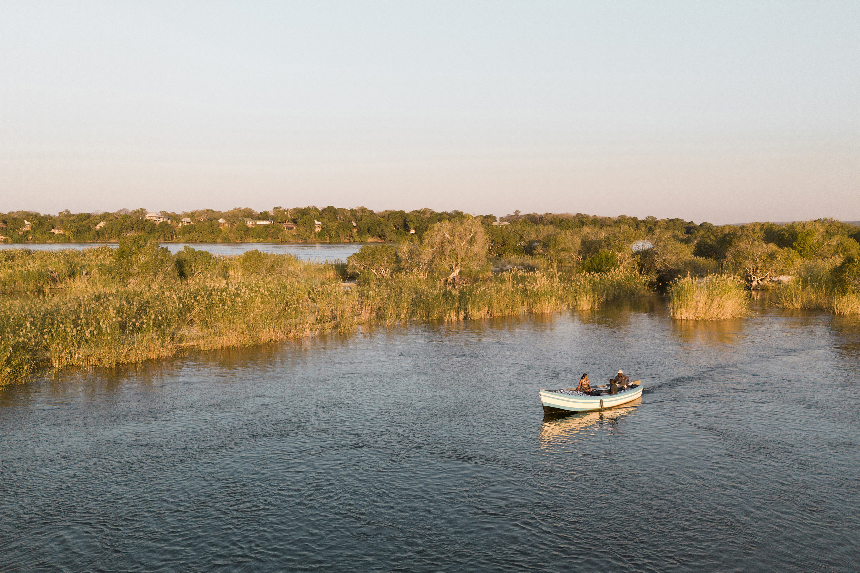 Two people in the electric dhow at Tongabezi Camp on the calm Zambezi River surrounded by lush greenery and reeds during sunset.