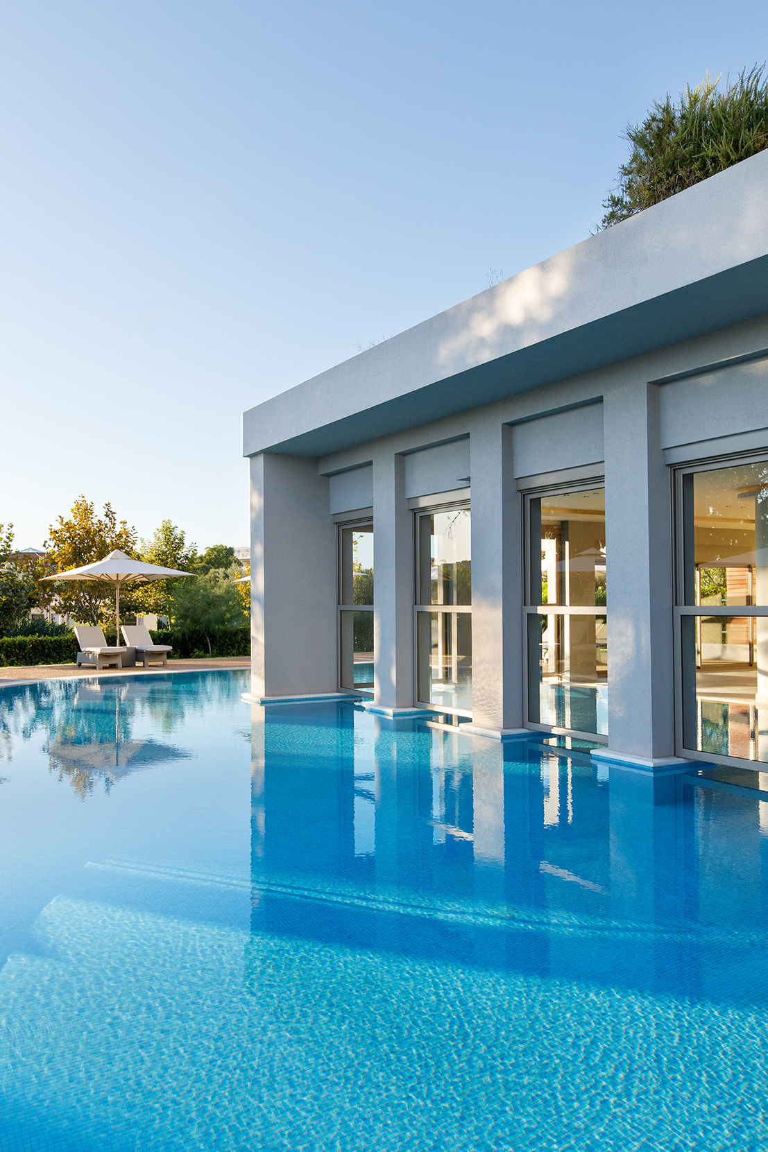 The large stone spa building at Ikos Olivia surrounded by a swimming pool with two sun loungers at the side under a clear sky