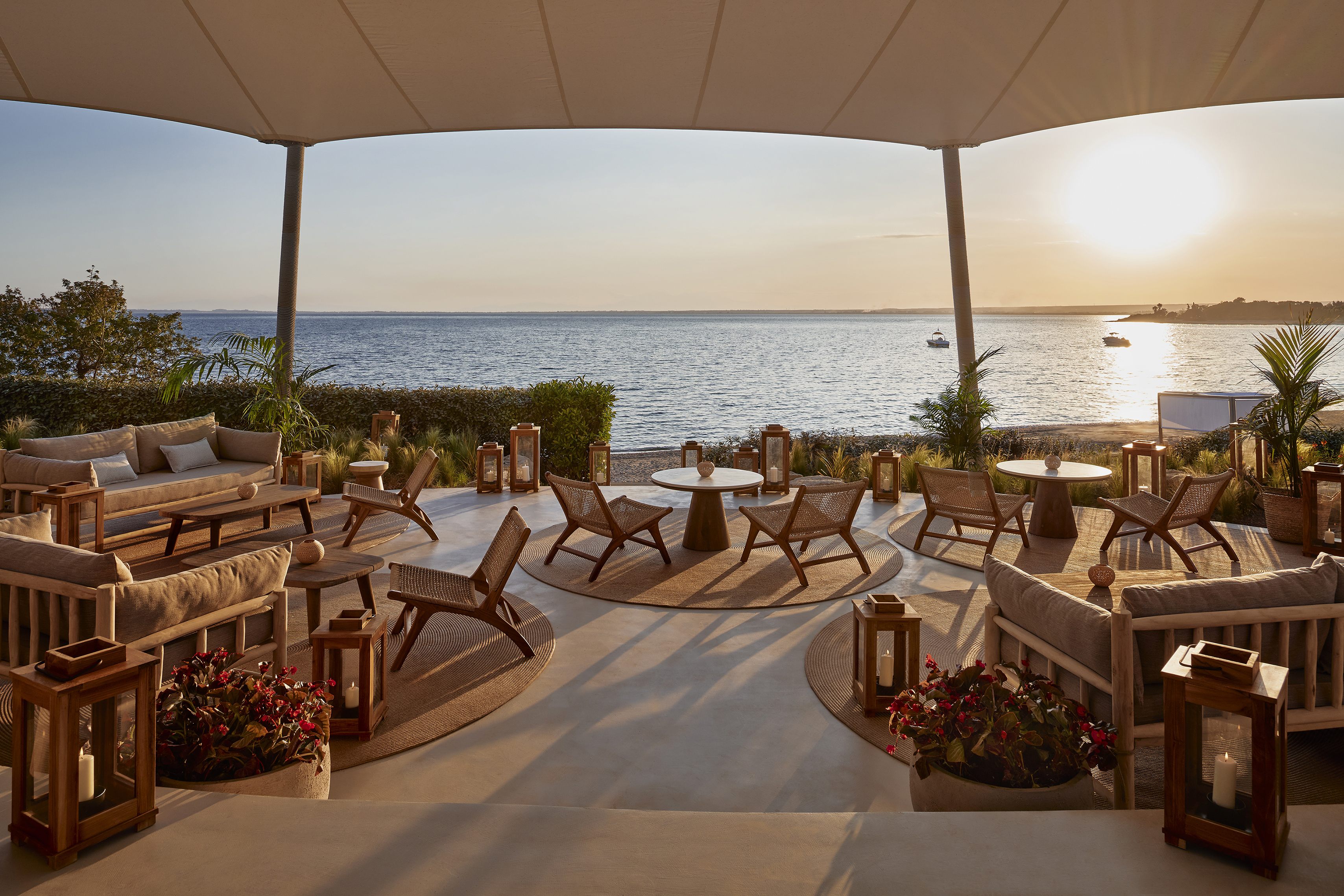 A large restaurant terrace with dining tables and sofas beneath a tented canopy by the side of the sea with boats in the distance
