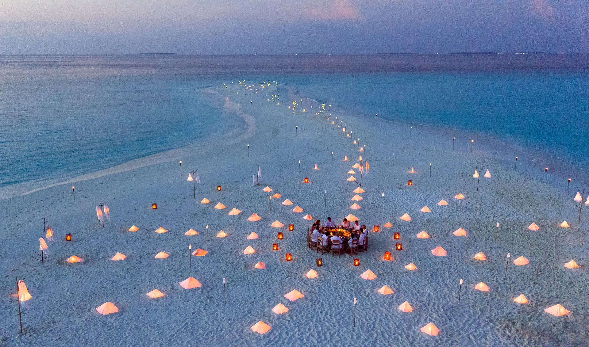 A bird's eye view of an evening dinner on the beach, people are gathered around a circular table and the sand is lined with lanterns