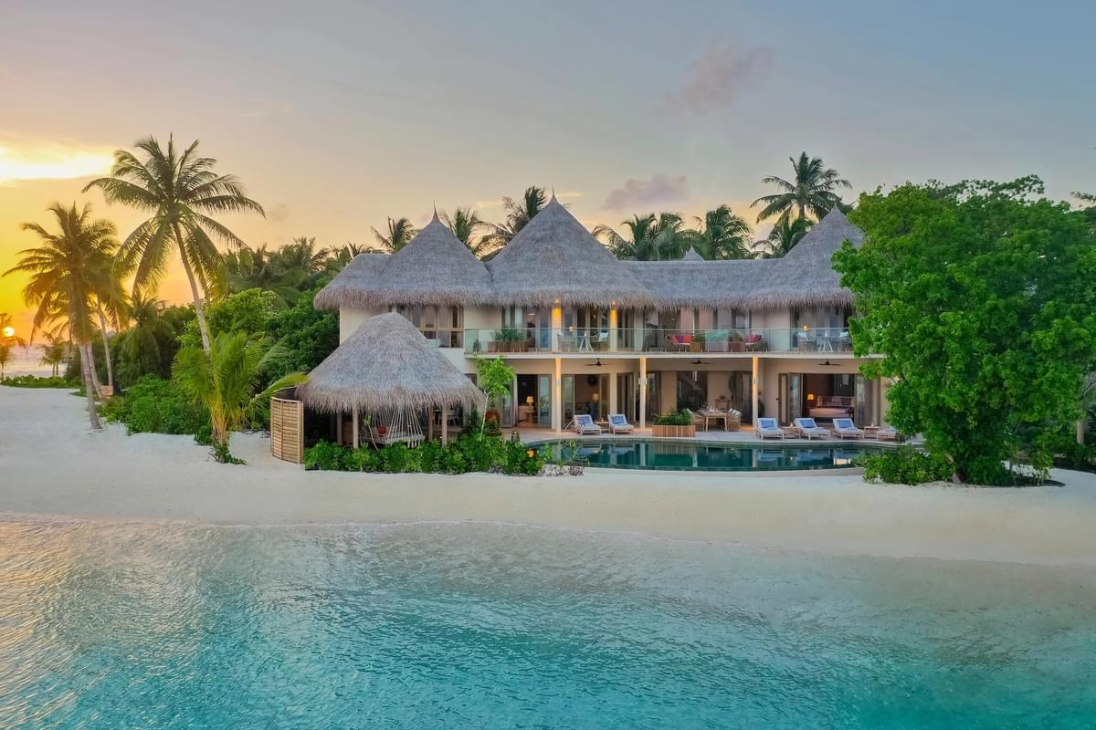 A luxurious beachfront villa with a thatched roof at sunset, surrounded by palm trees and clear skies.